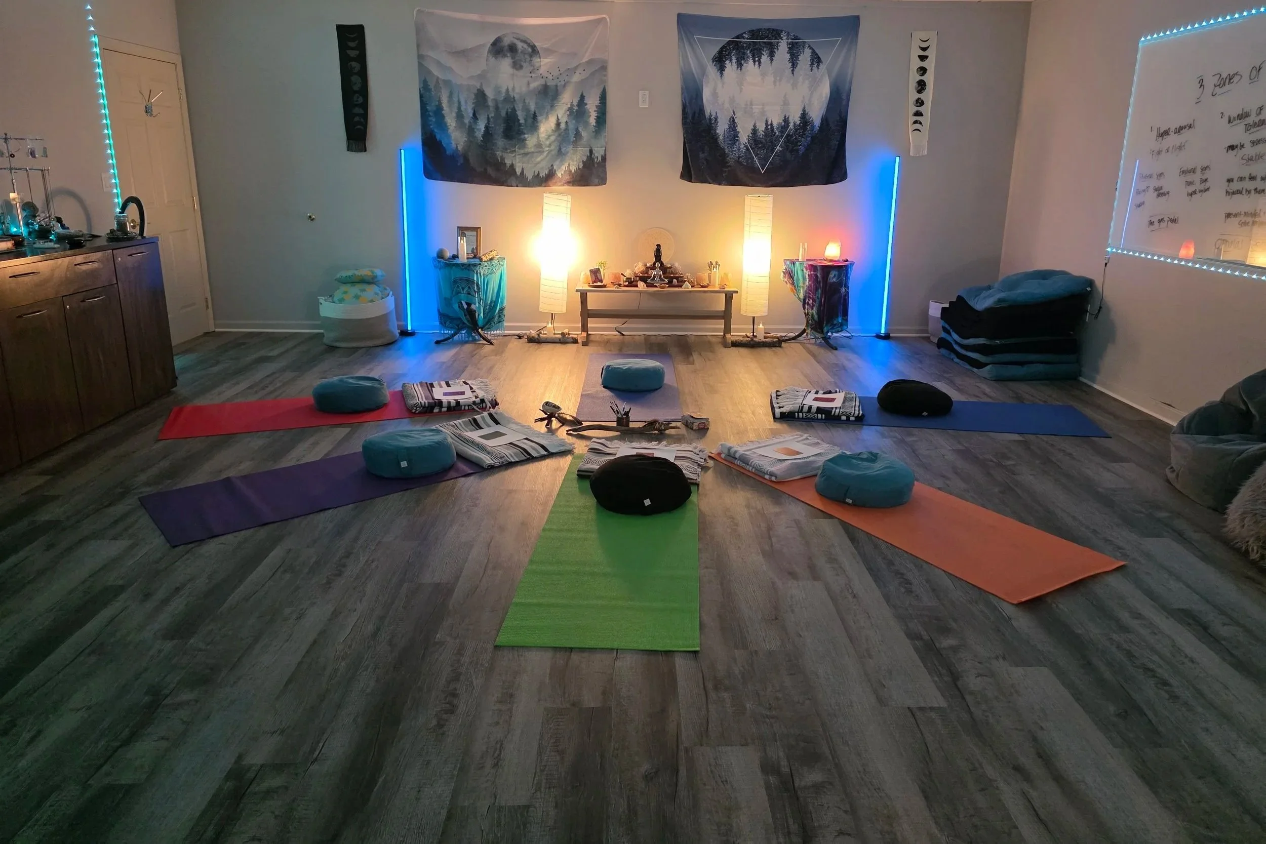 A meditation or yoga room set up with eight colorful mats and cushions arranged in a circle. The room features wall tapestries with forest and moon images, ambient lighting, and a small altar at the back with candles and spiritual items.