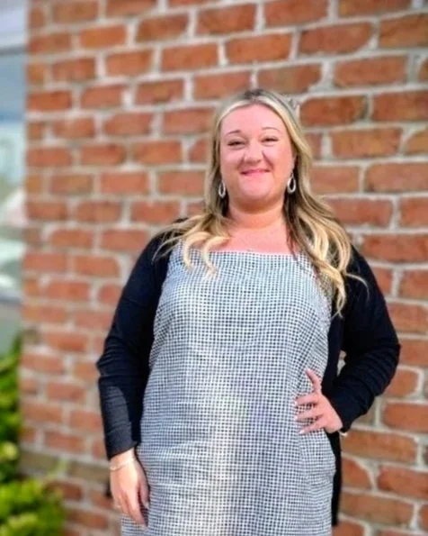 A woman with blonde hair smiling outdoors, standing in front of a brick wall, wearing a patterned dress and a dark cardigan.