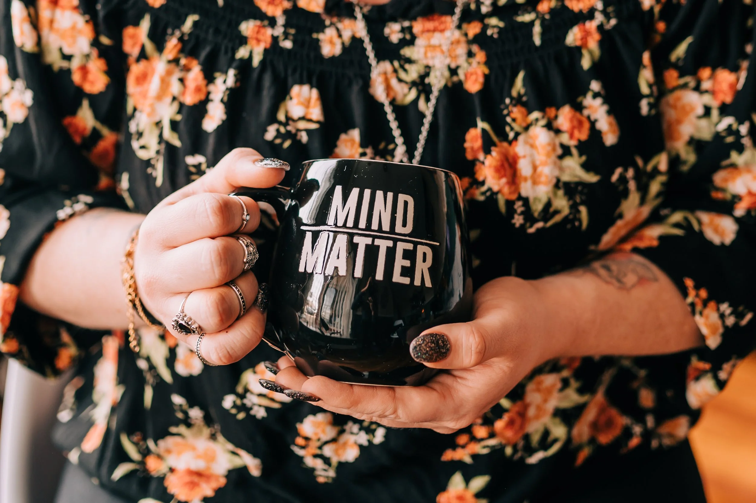 Person holding a black mug with the words "MIND MATTER" in white lettering, wearing a black floral blouse and multiple rings and bracelets.