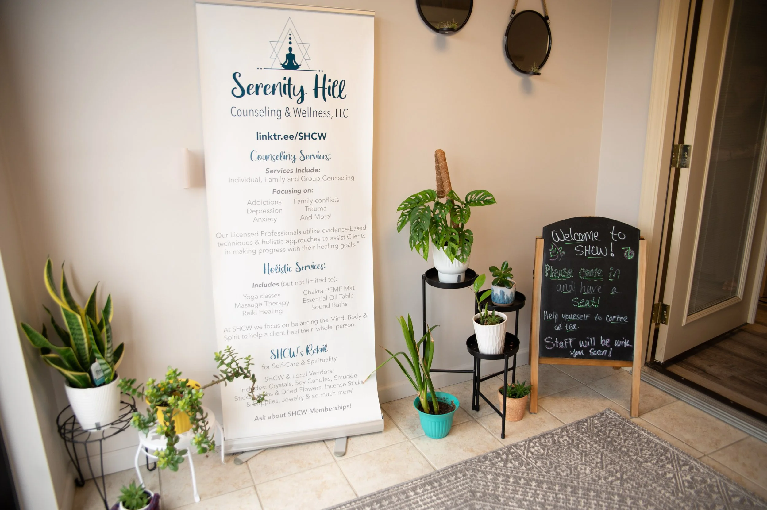 Indoor entrance area with a board sign for Serenity Hill Counseling & Wellness LLC, a chalkboard sign welcoming visitors to a show, and several potted plants near the wall.