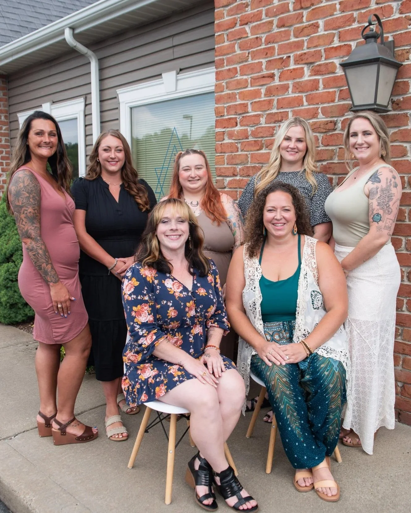 Group of eight women smiling outdoors, some sitting and some standing, in front of a brick wall and house exterior.