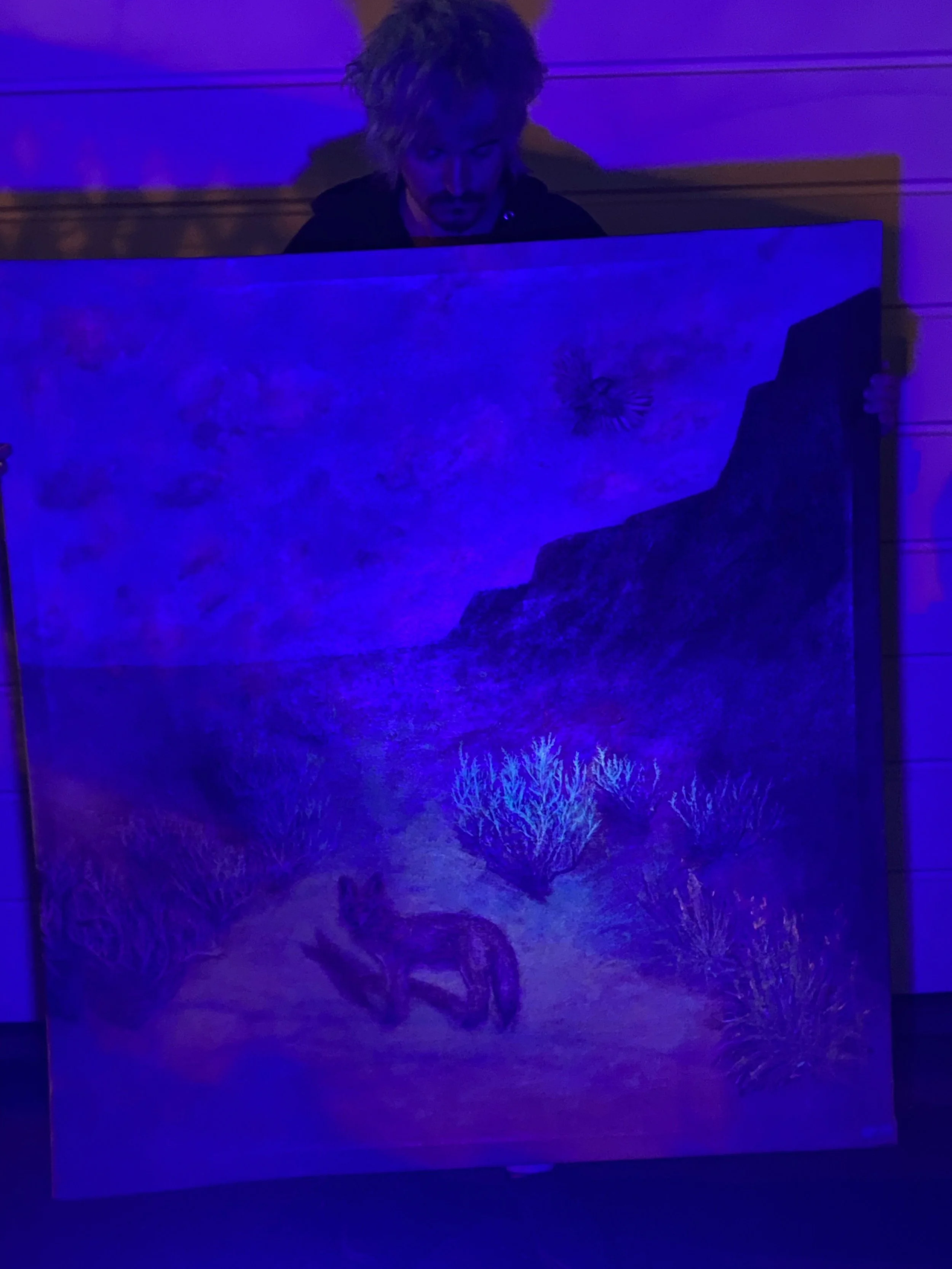 Person with light-colored hair holding a painting of a desert landscape with cacti, rocks, and a canyon, illuminated under purple lighting.