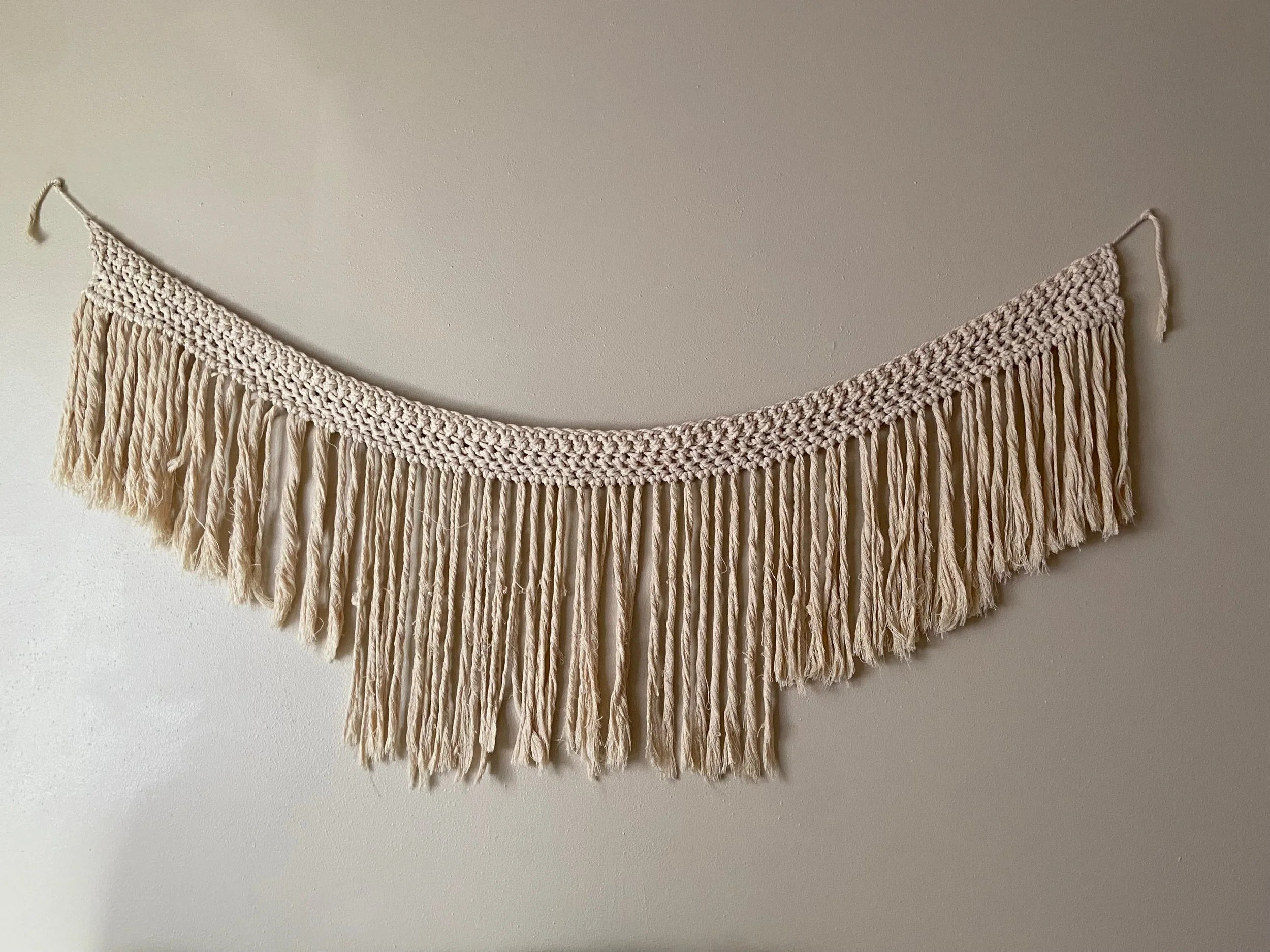 A decorative wall hanging with a crocheted top and long fringes below, suspended on a beige wall.