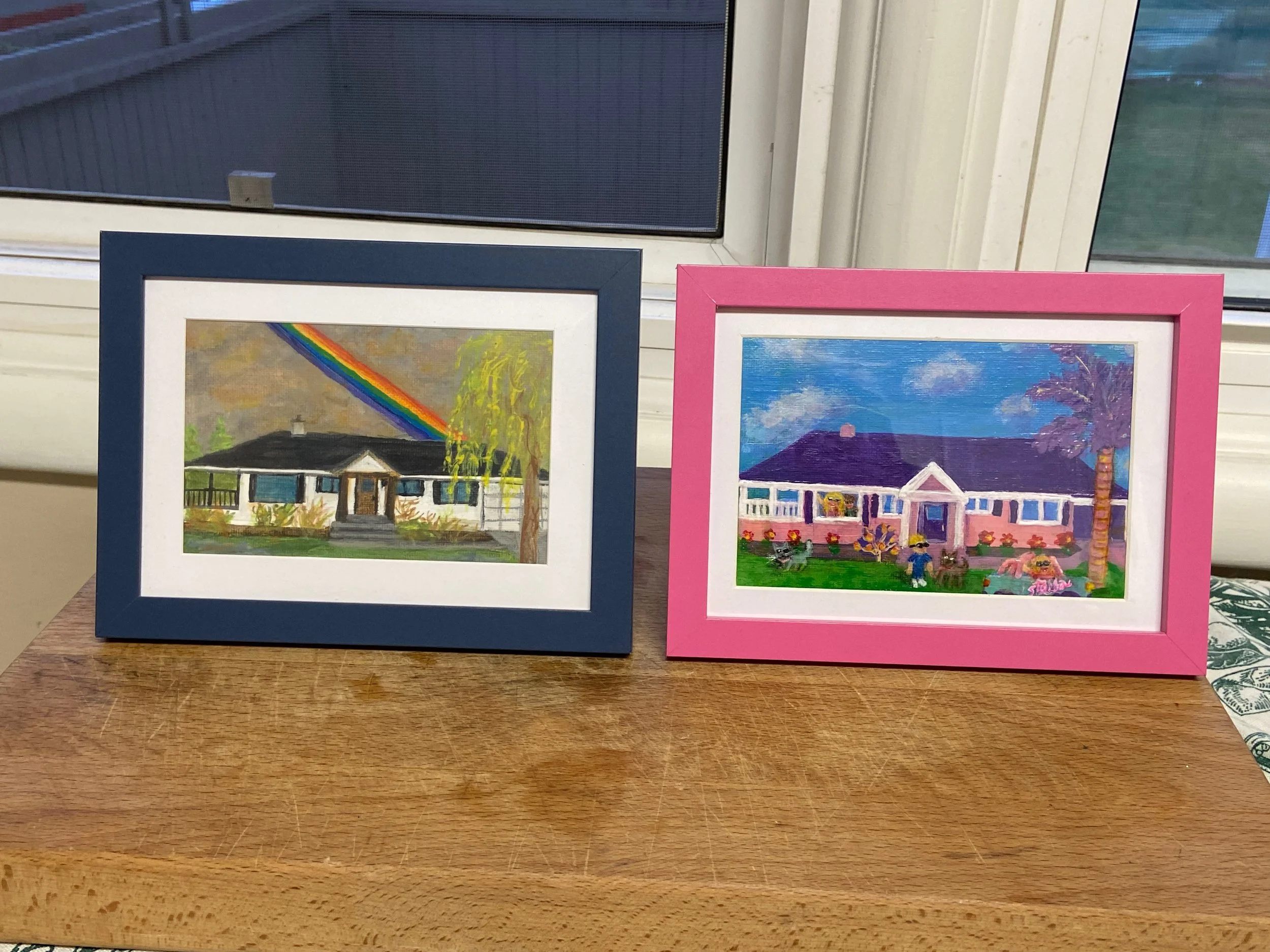 Two framed watercolor paintings on a wooden table; one with a house and rainbow, the other with a pink house and garden scene.