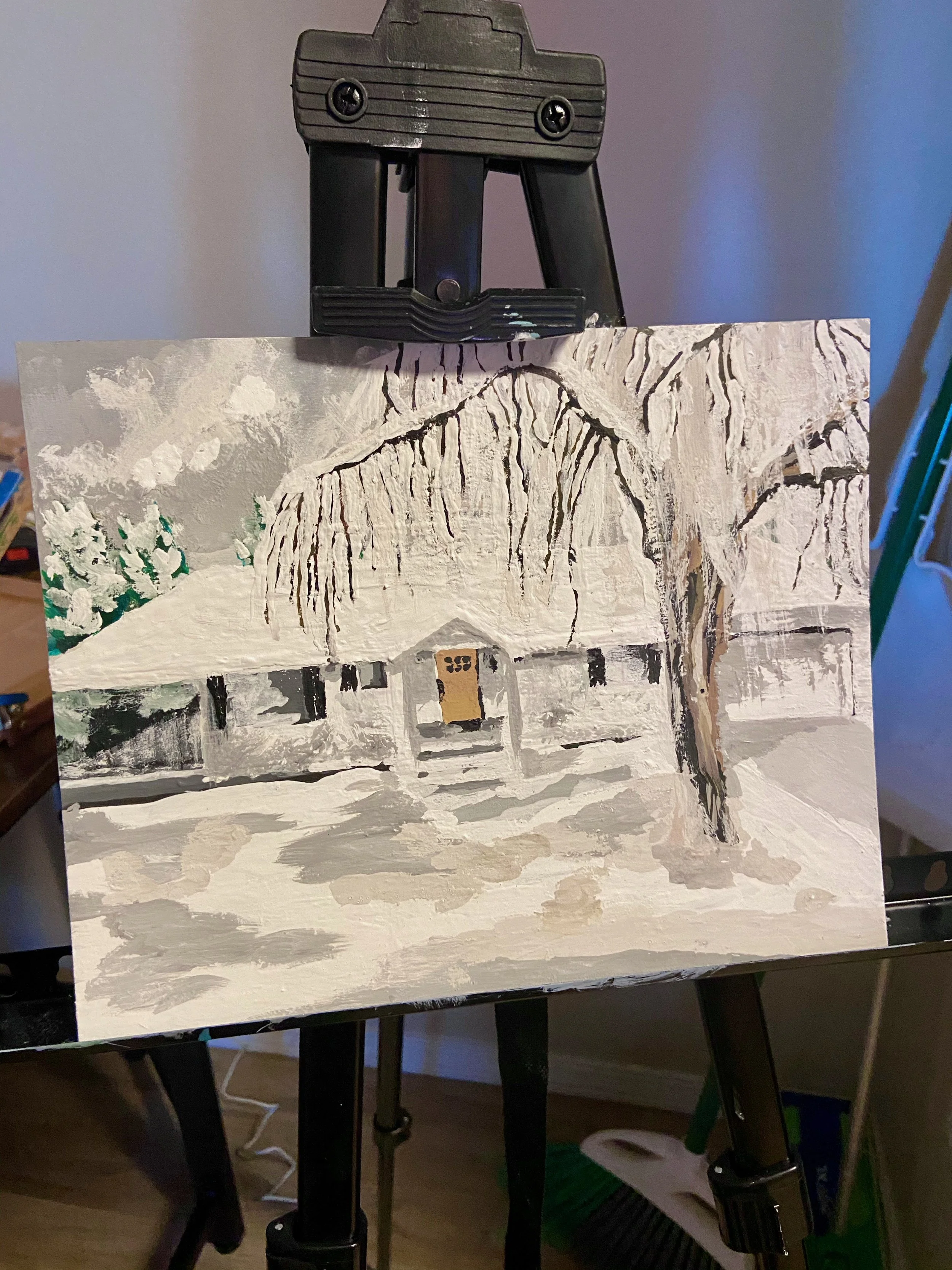 A painting of a snow-covered house with a thatched roof, surrounded by snow and trees, on an art easel.
