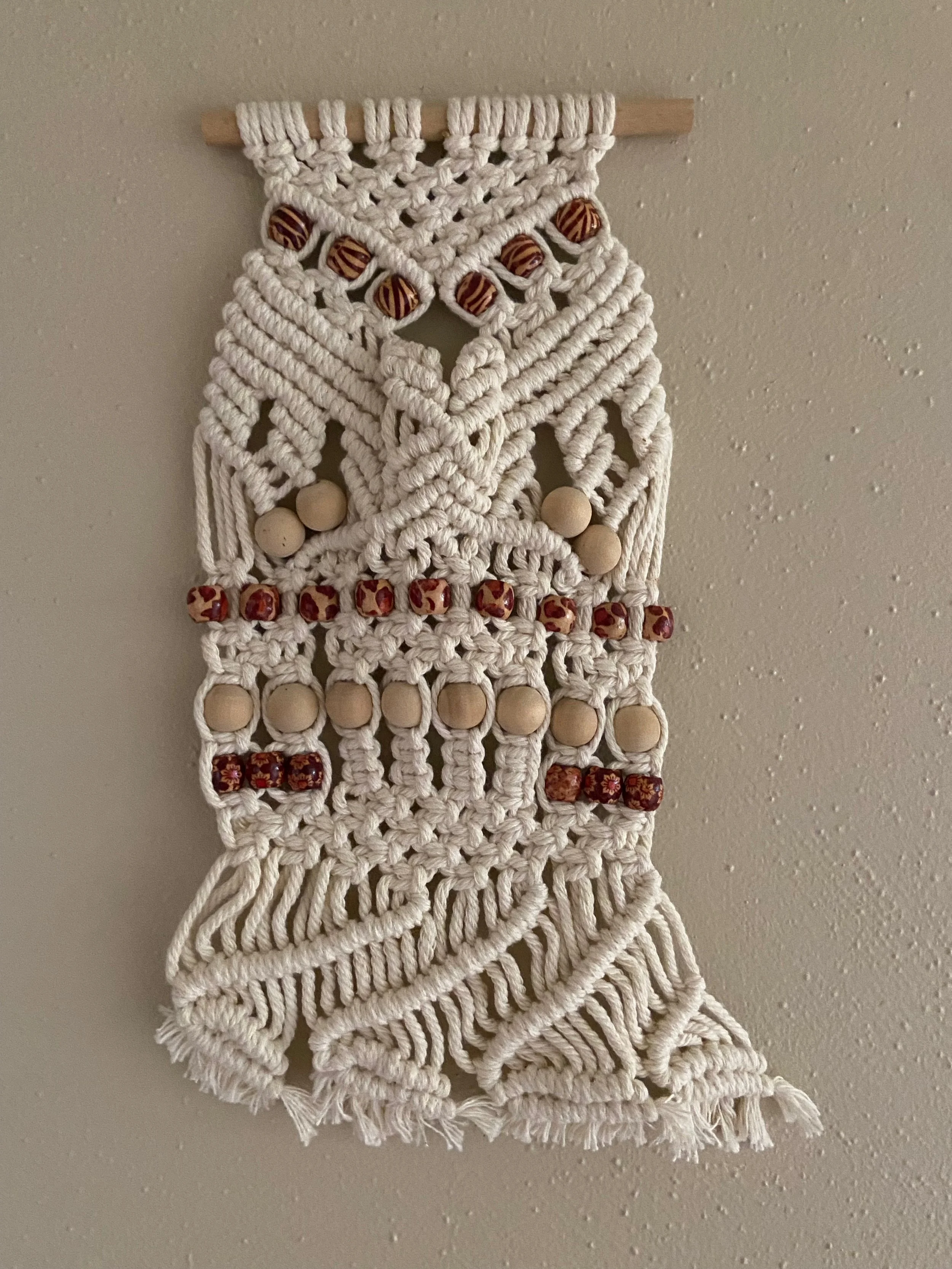 A decorative wall hanging made of white macrame with wooden beads and small fabric beads.