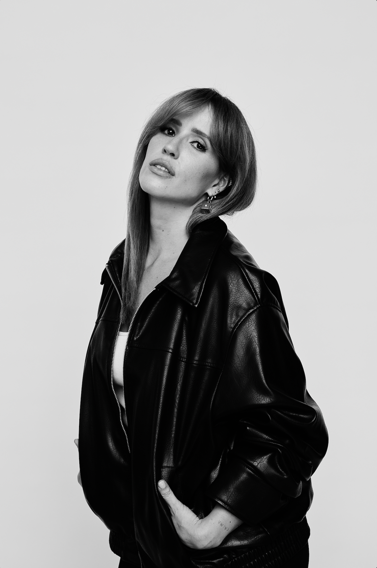 Retrato de Marcela Ceribelli com cabelos longos e lisos, usando jaqueta de couro preta, com acessórios de piercing na orelha, posando de lado com expressão confiante.