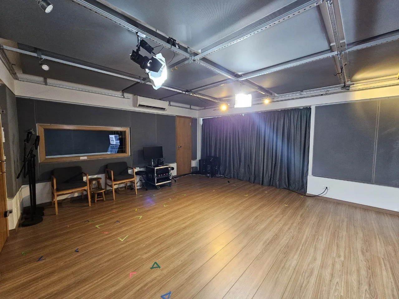 Sala de gravação com piso de madeira, paredes acústicas, cortinas negras, equipamentos de iluminação, câmeras de vídeo, e cadeiras.