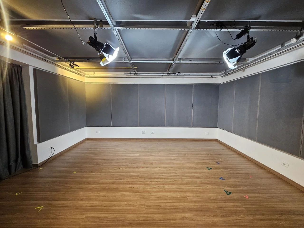 Sala de gravação vazia com piso de madeira, paredes acusticas cinzas e iluminação de estúdio com iluminação em braco, em um ambiente fechado.