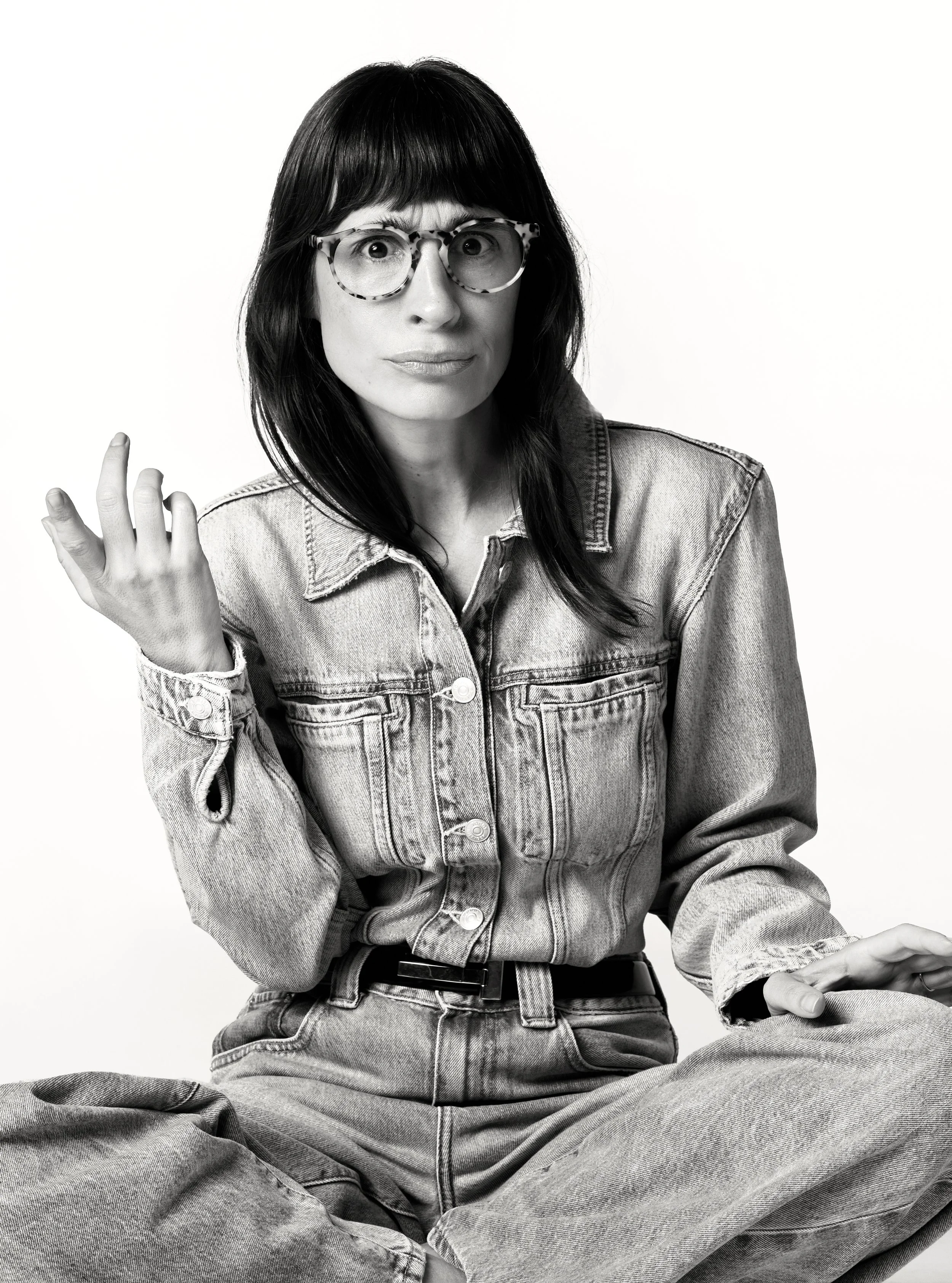 Camila Fremder de cabelo escuro e óculos de armação tartarugada, vestindo jaqueta jeans, sentada com pernas cruzadas, em fundo branco