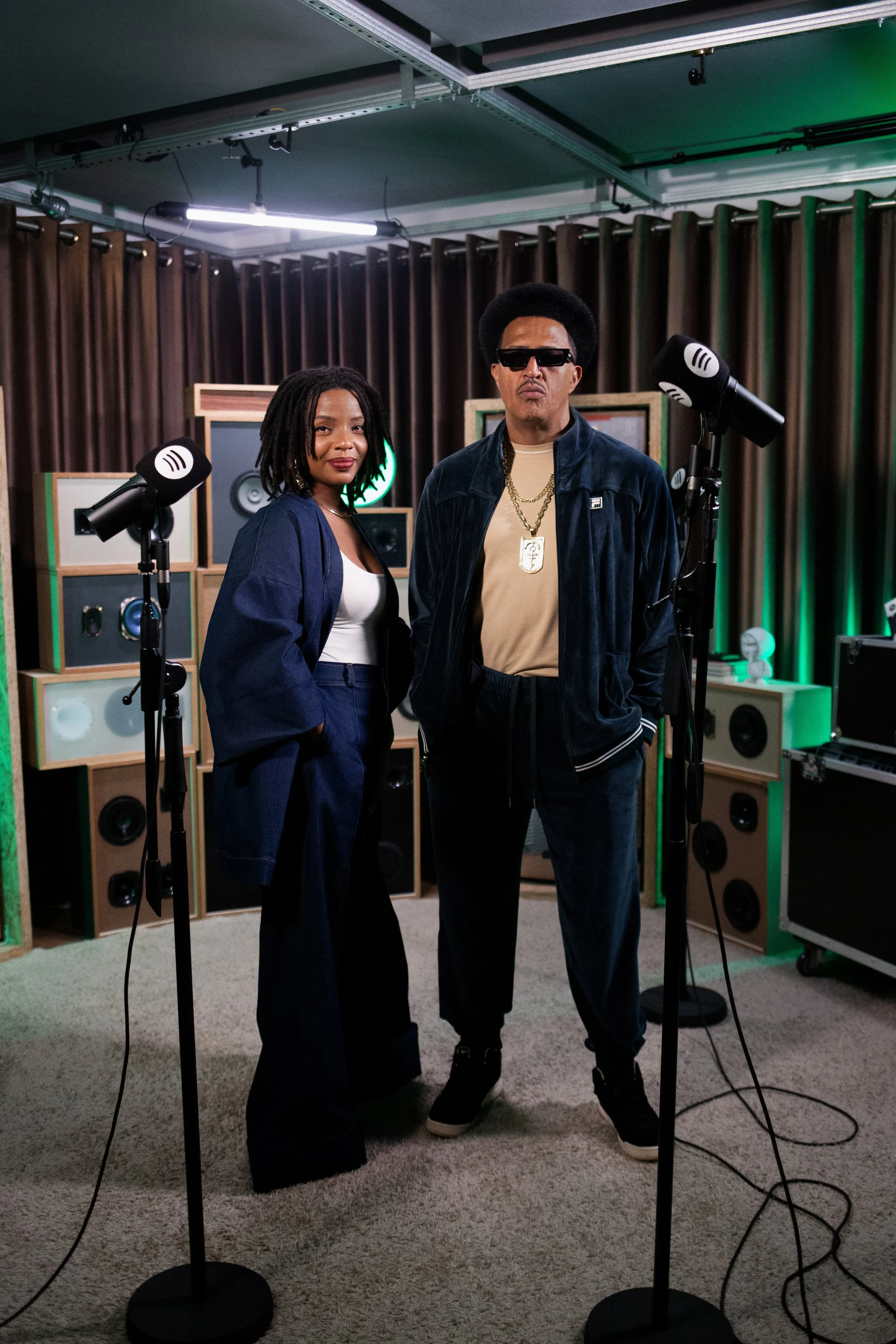 Mano Brown e Semayat Oliveira posando em estúdio de gravação do Mano a Mano com alto-falantes e equipamento de som ao fundo, roupas estilosas e expressão confiante.