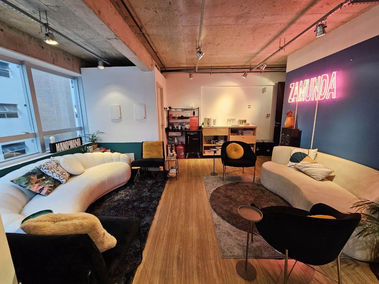 Sala de estar com sofá branco curvo, poltronas pretas, uma parede azul com neon rosa escrito 'ZAMUNDA', janelas grandes, piso de madeira, iluminação moderna, decoração com almofadas coloridas e plantas.