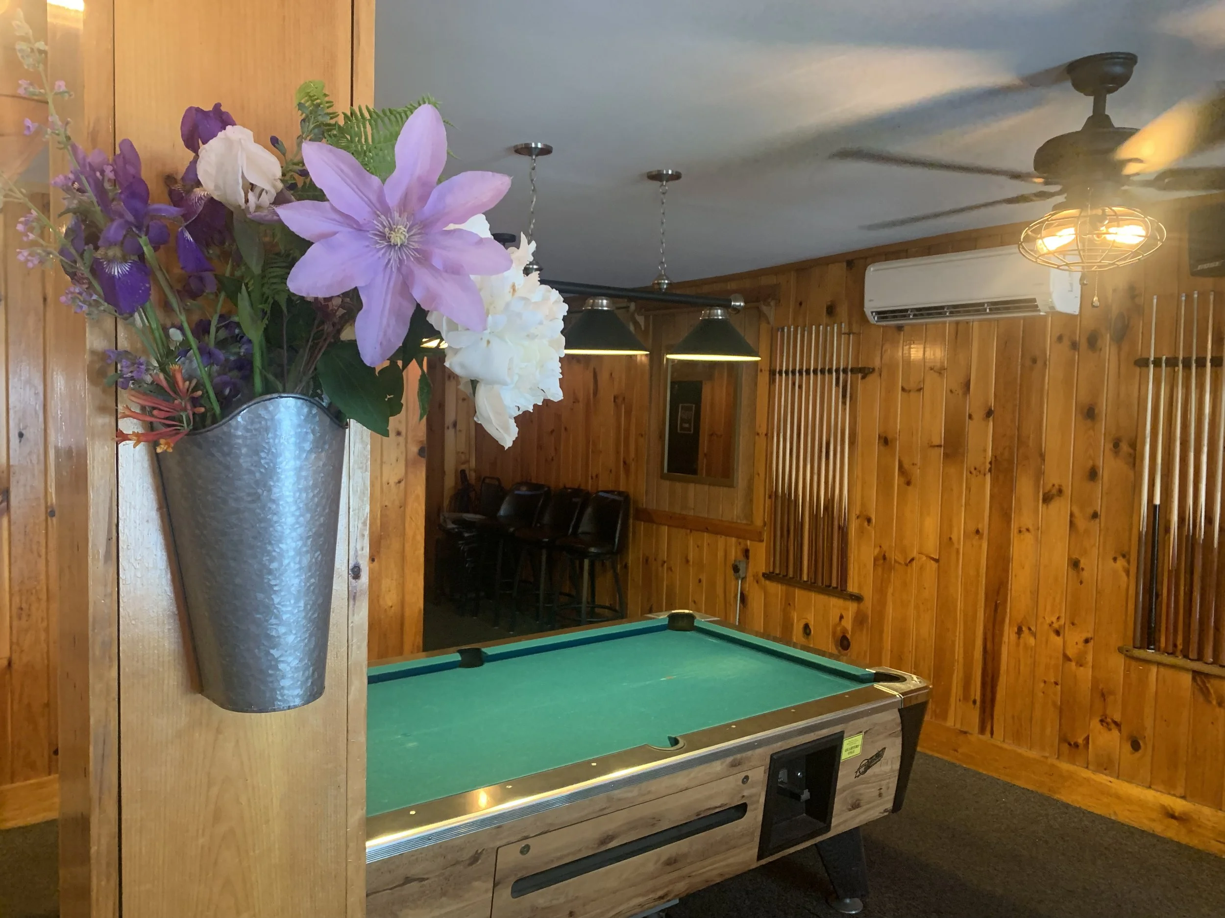 A metal vase with purple, purple and white flowers attached to a wooden wall in a game room. The room has wooden walls, a pool table, hanging lights, an air conditioning unit, ceiling fan, and stacked chairs in the background.