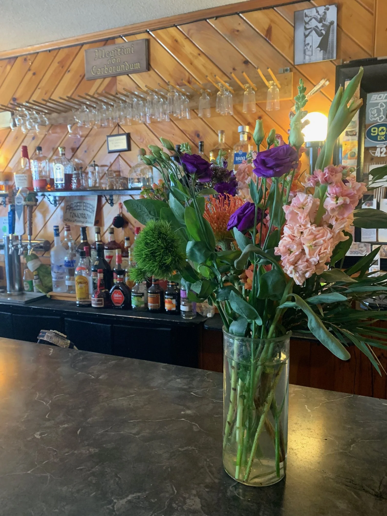 A glass vase with a colorful arrangement of flowers including purple lisianthus, pink stock, green fern, and orange protea, placed on a marble countertop in a bar with wooden paneling and various liquor bottles in the background.