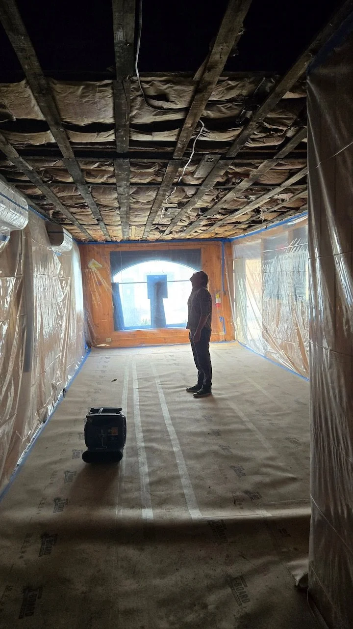 Person standing in a room under construction with exposed ceiling beams and insulation, plastic sheeting covering the walls, and a window at the far end.