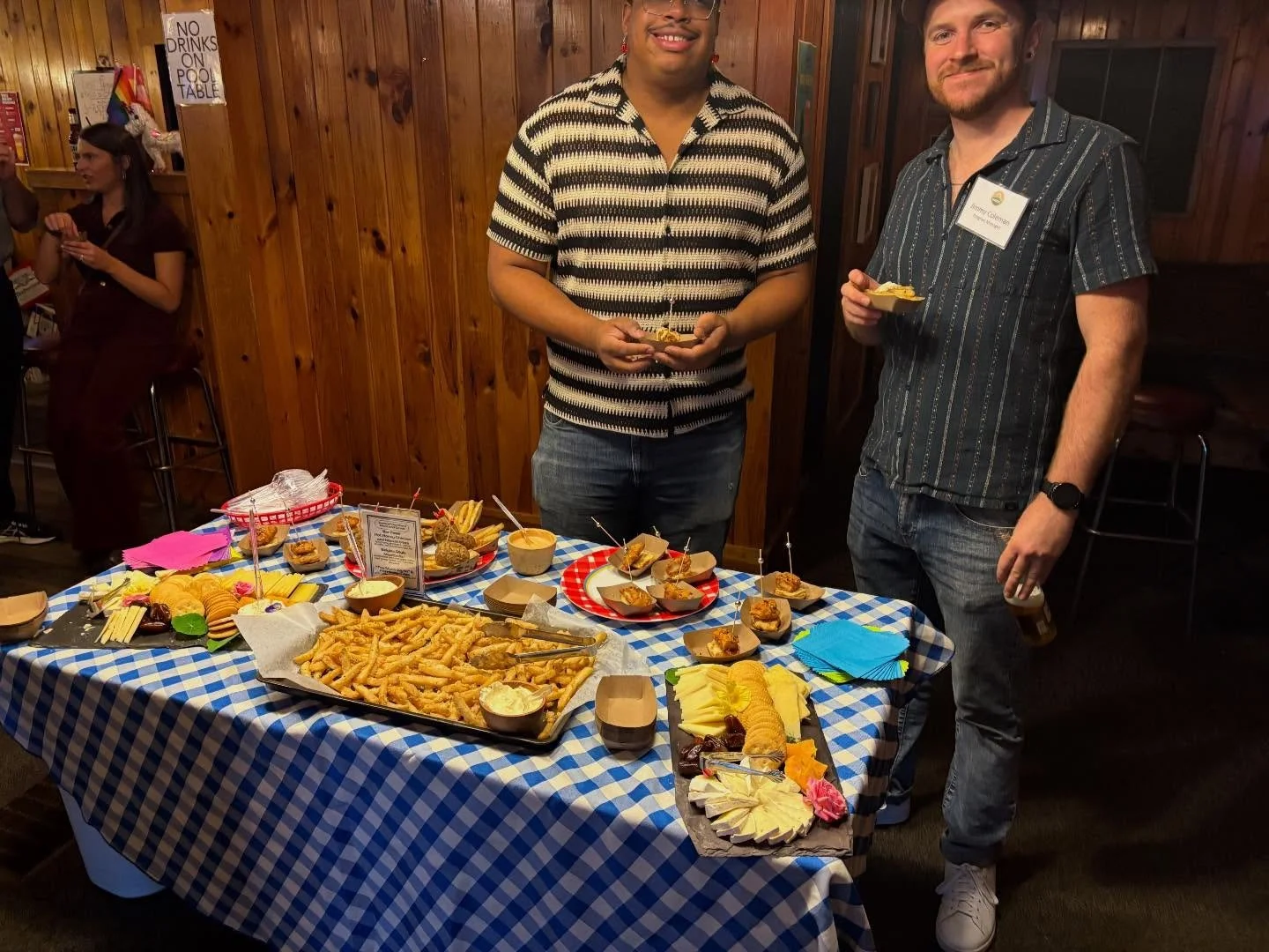 Two men standing at a table with appetizers in a social setting, with a woman in the background, wood-paneled walls, and a sign that reads "No drinks on pool table."