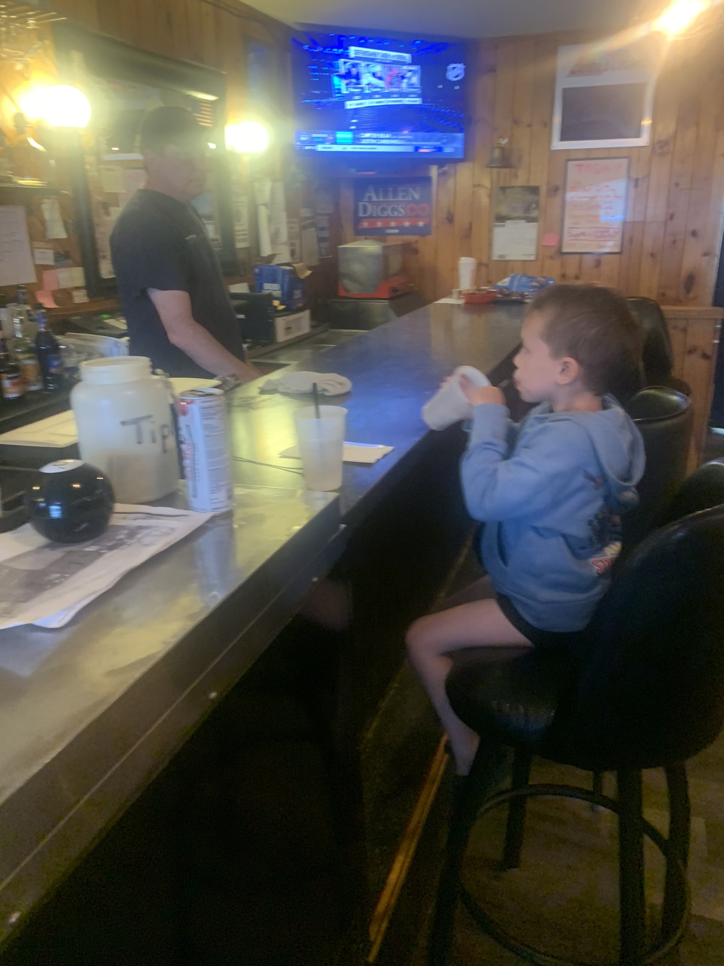 A young boy sitting at a bar counter drinking from a cup inside a rustic-style bar with wooden walls, a television on the wall, and various items such as a tip jar and condiments on the counter.