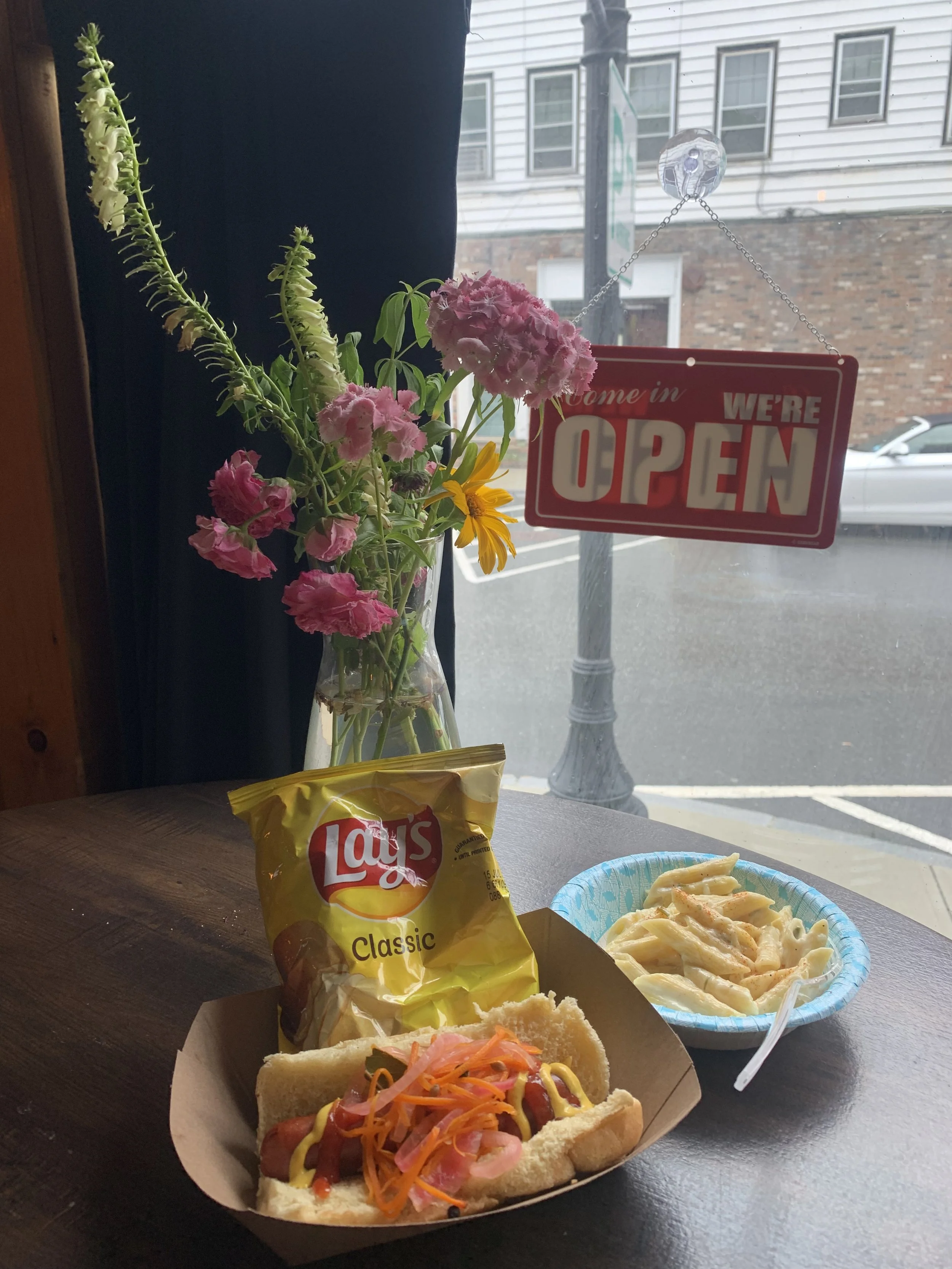 A table inside a restaurant with a hot dog topped with mustard, ketchup, and pickled vegetables, a bag of Lay's Classic potato chips, and a bowl of macaroni and cheese. There is a vase with pink and yellow flowers and a red 'We're Open' sign hanging 