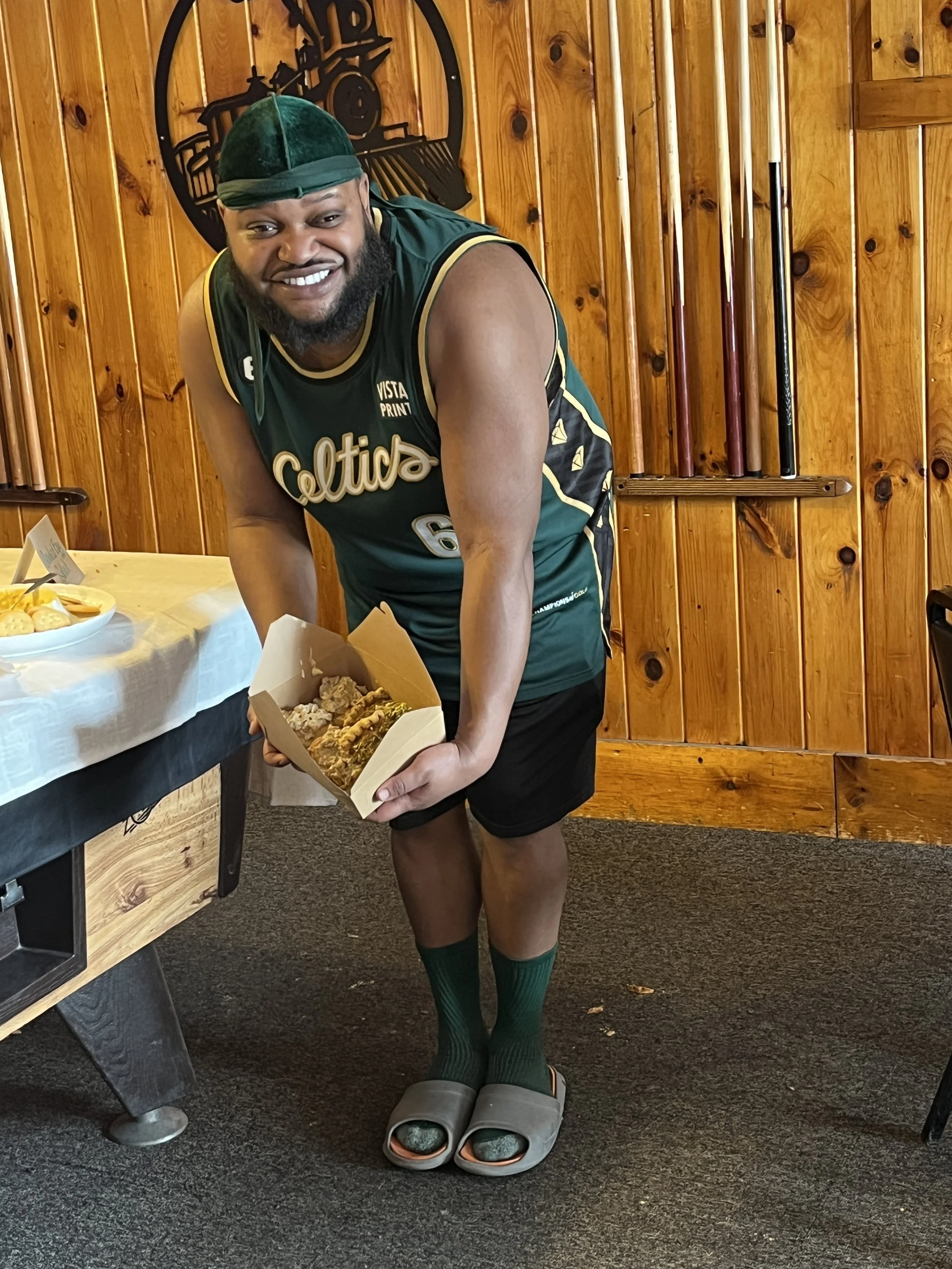 A smiling man wearing a green sports jersey and hat, black shorts, green socks, and gray slides, holding a cardboard container of food, standing indoors in front of a wooden wall with a paddle rack and a clock.