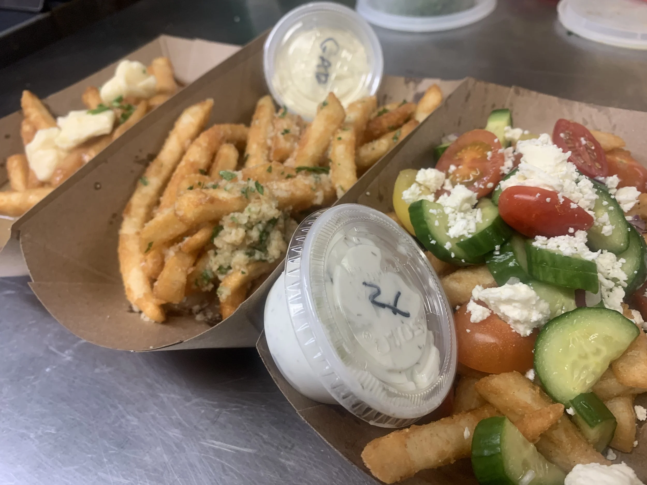 Crispy French fries served with small containers of ranch dressing, topped with herbs, and a side salad with cherry tomatoes, sliced cucumbers, green onions, and crumbled feta cheese.