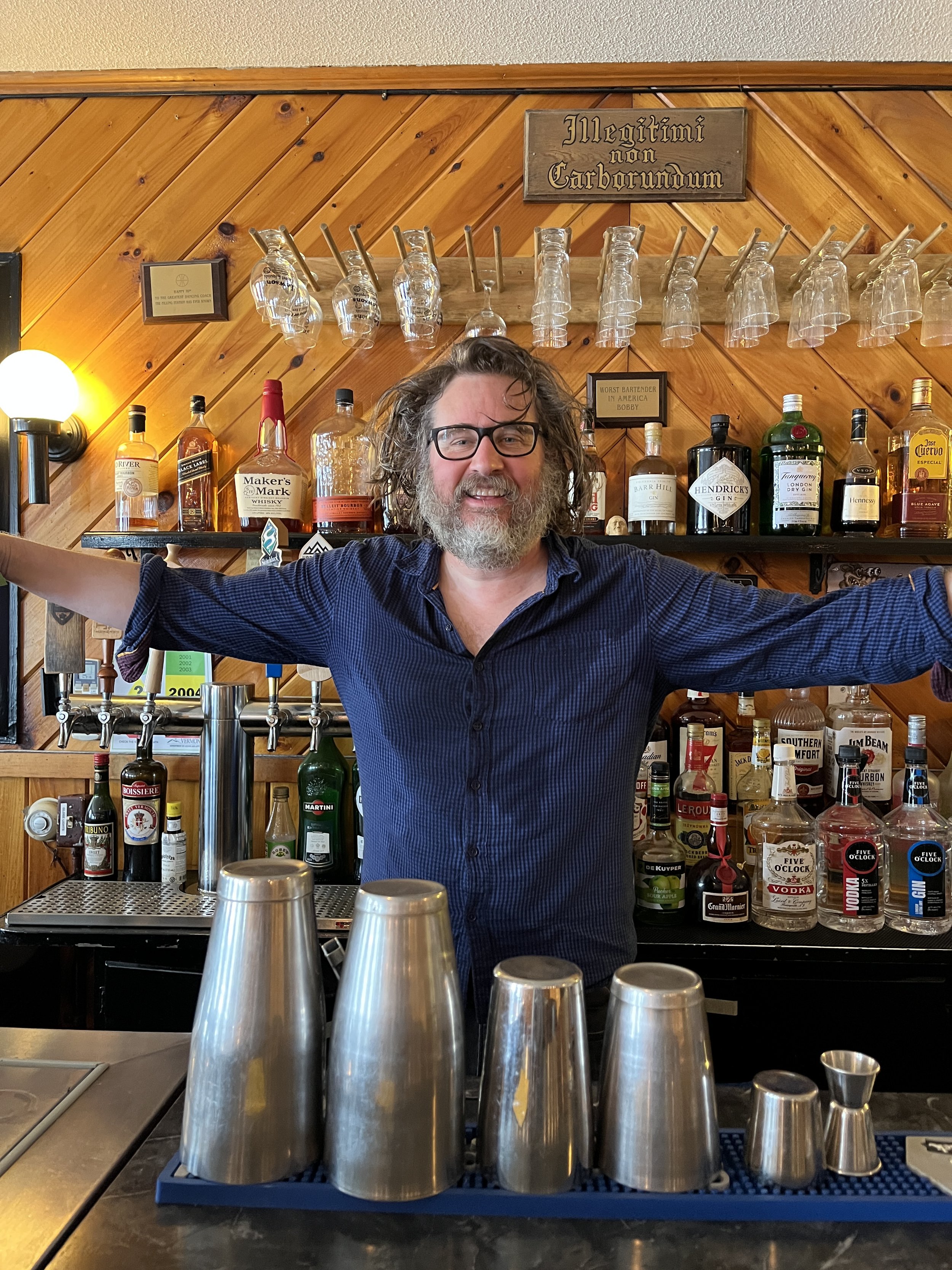 A smiling bartender with glasses and long hair standing behind a bar with arms out wide, in front of a shelf of various liquor bottles and upside-down glassware.