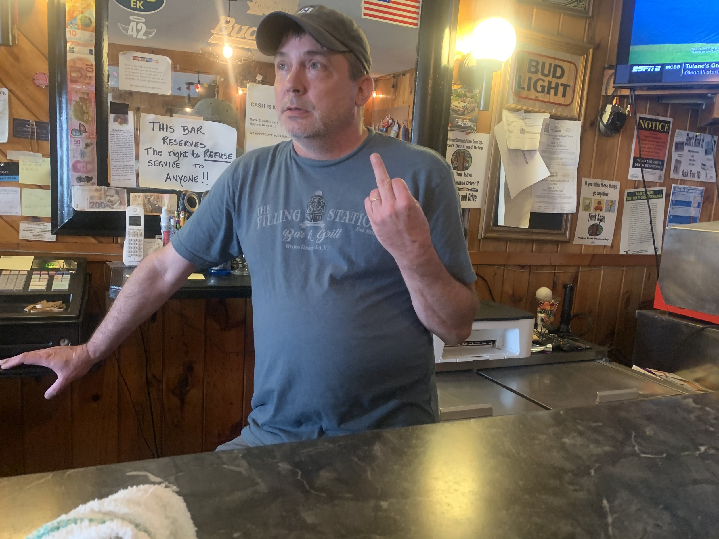 A man standing behind a bar counter gives the middle finger. He is wearing a gray T-shirt and a gray cap inside a wooden-paneled bar or restaurant. Various signs and notices are posted on the walls behind him.