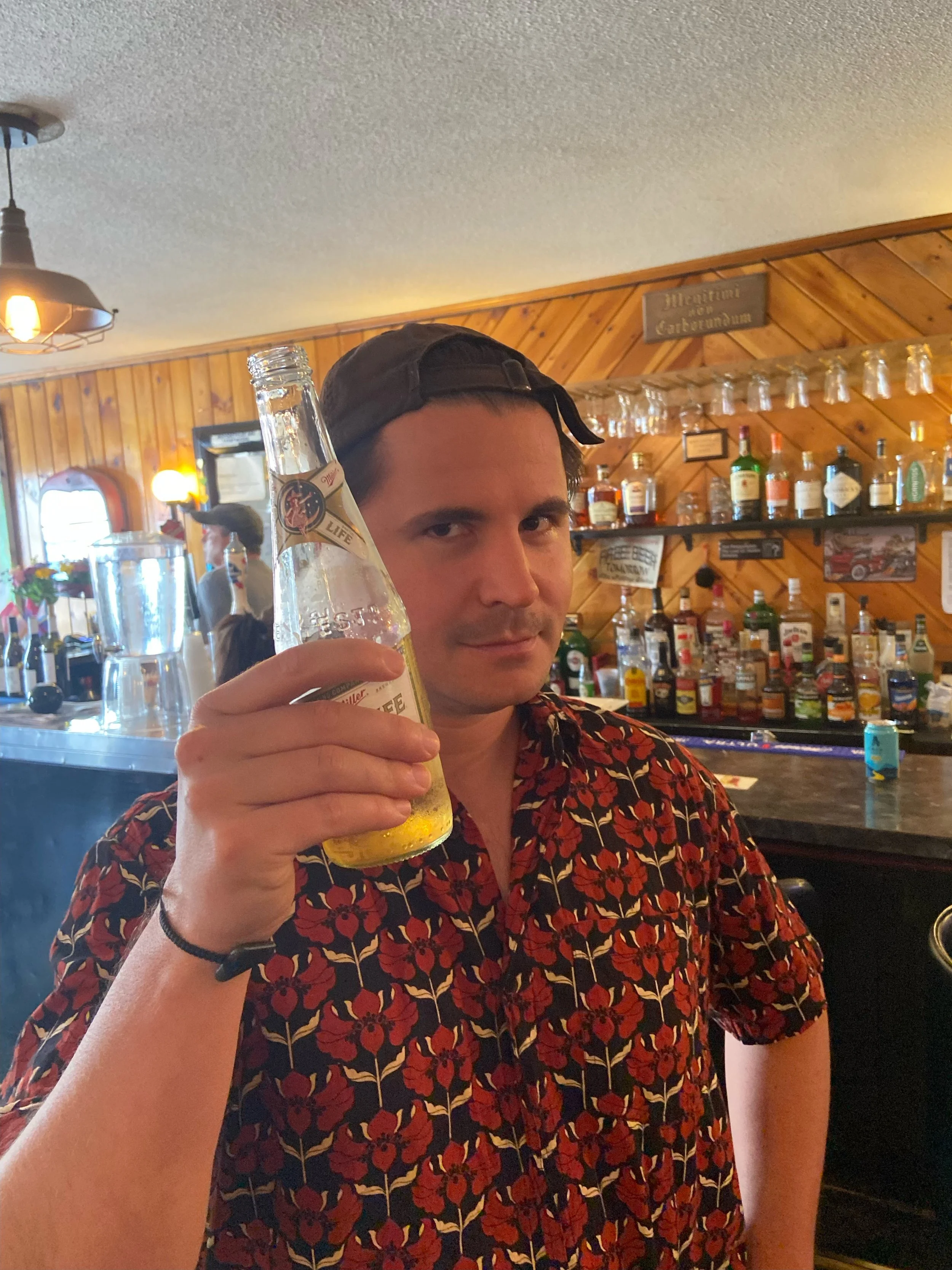 A man wearing a black cap backwards and a floral red and black shirt holds up a clear glass bottle of beer in a bar with wooden walls and a variety of liquor bottles on shelves behind him.