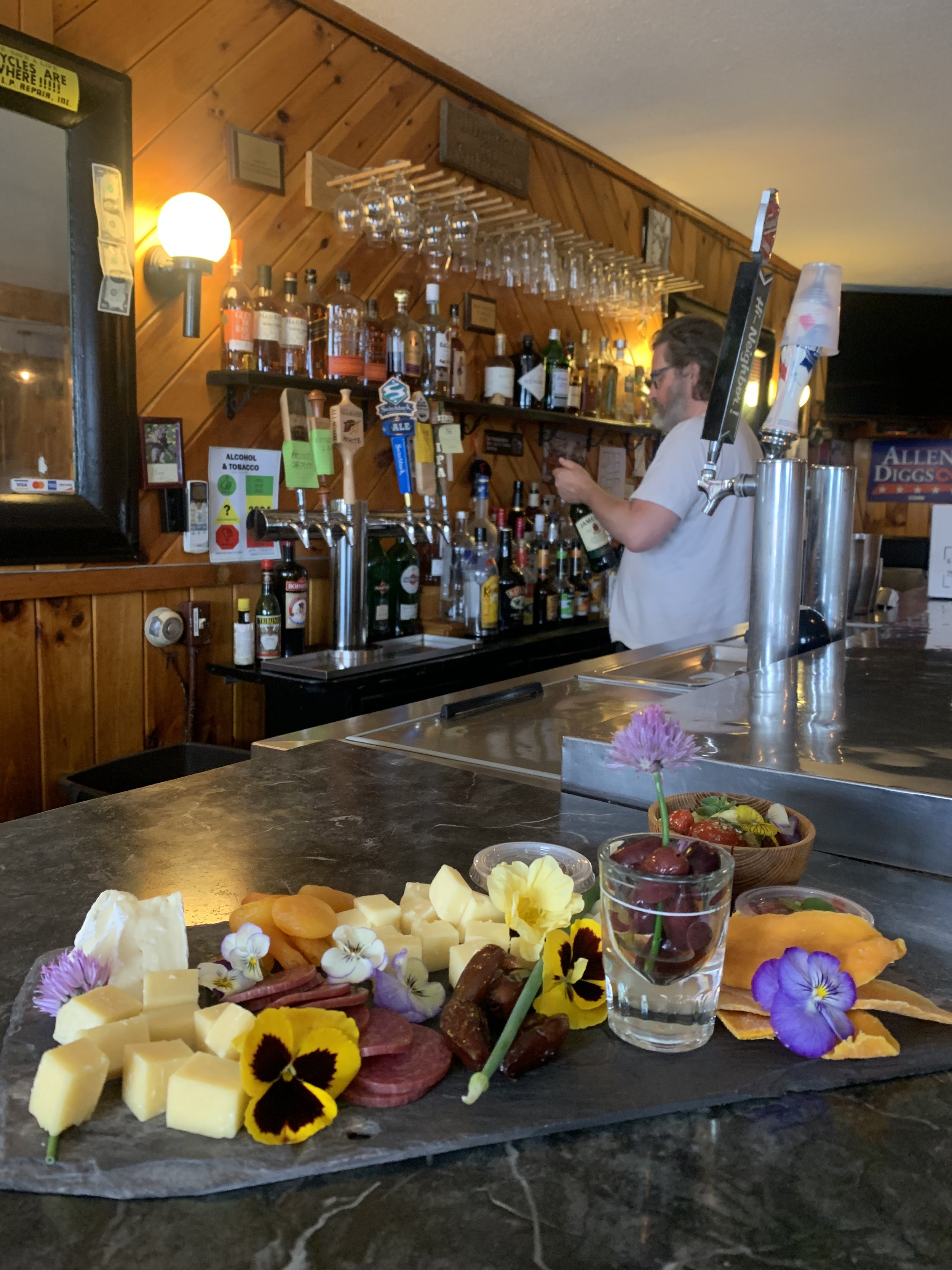 A bar with a display of assorted cheeses, meats, fruits, and edible flowers on a slate board, and a bartender behind the counter pouring a drink, with liquor bottles and glassware on shelves in the background.