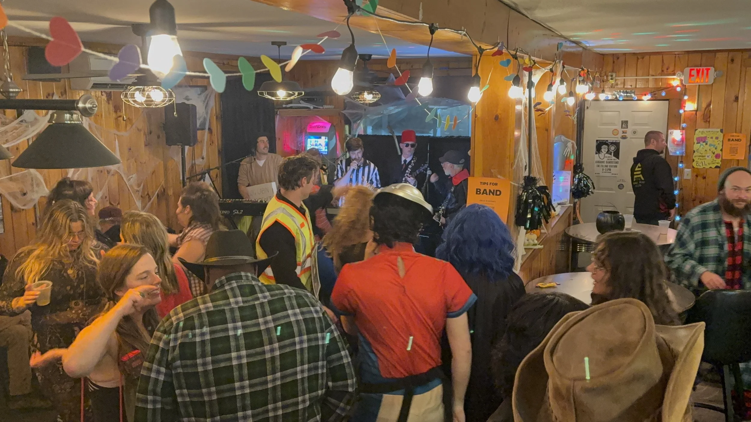 People at a Halloween party with costumes, dancing, and a band playing on stage in a decorated room with spider webs and string lights.