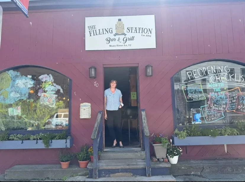 A woman standing at the entrance of The Filling Station Bar & Grill in White River Junction, Vermont. The building has a reddish-brown exterior with a sign above the door, and large windows displaying colorful signs and decorations. There are potted plants on the steps leading up to the entrance.