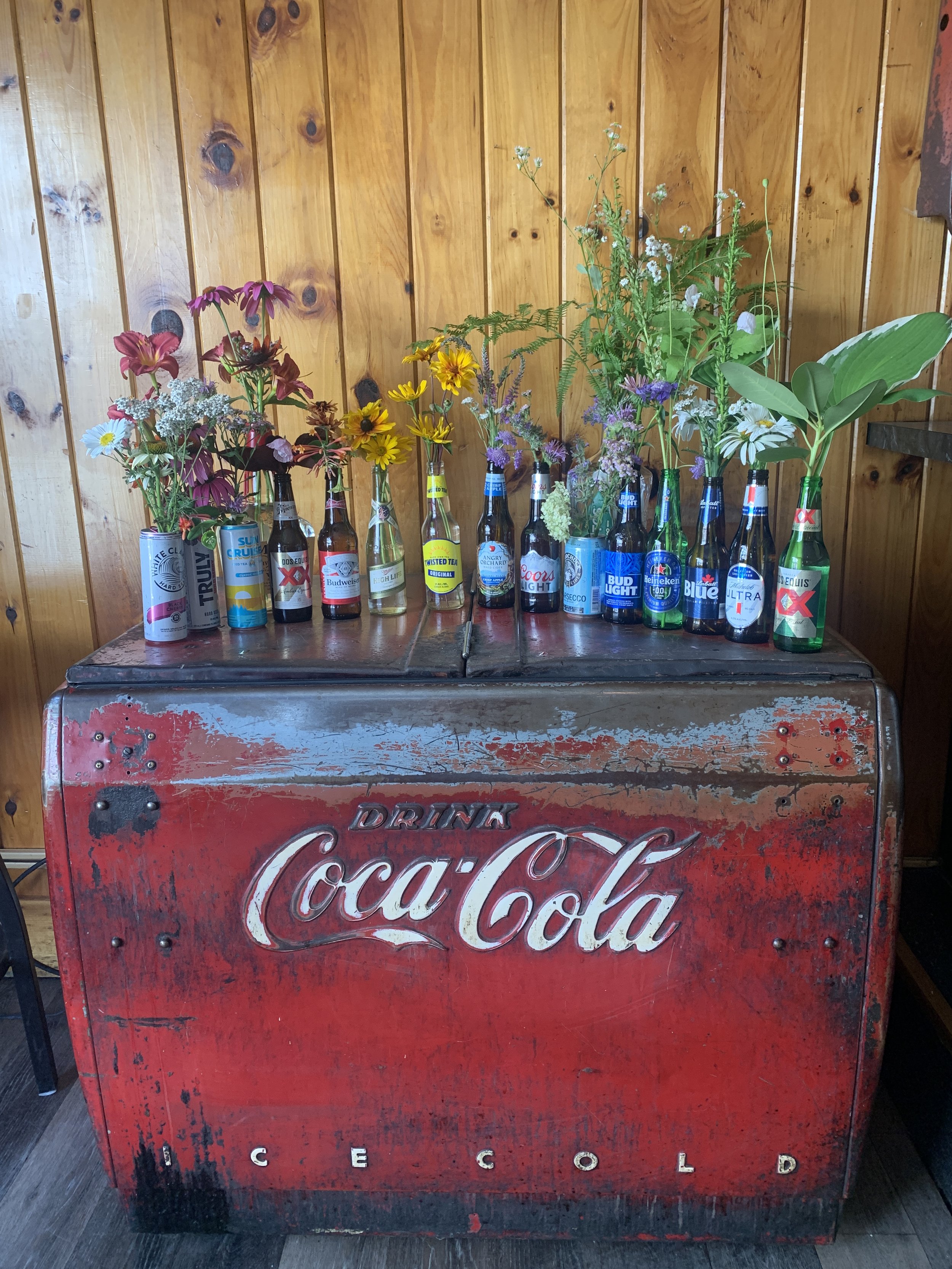 An old, red Coca-Cola ice cooler with a weathered and distressed appearance, topped with various cans and bottles of beer and soft drinks, each holding flowers in different arrangements, against a wooden wall background.