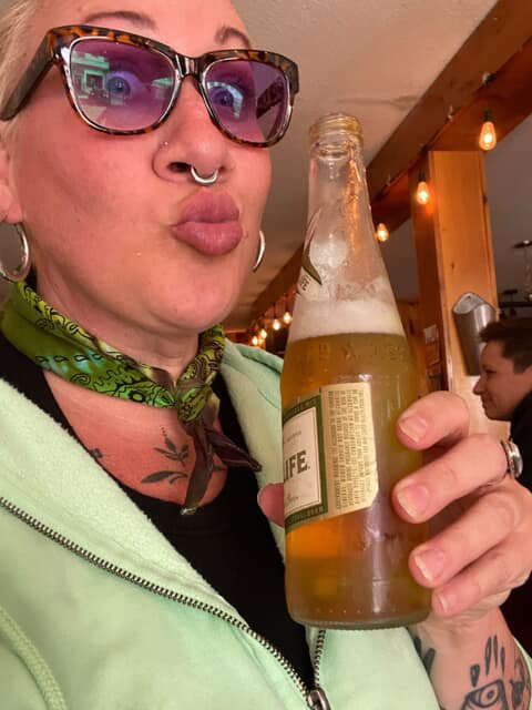 Person with glasses and a nose ring holding a beer bottle inside a bar or restaurant.