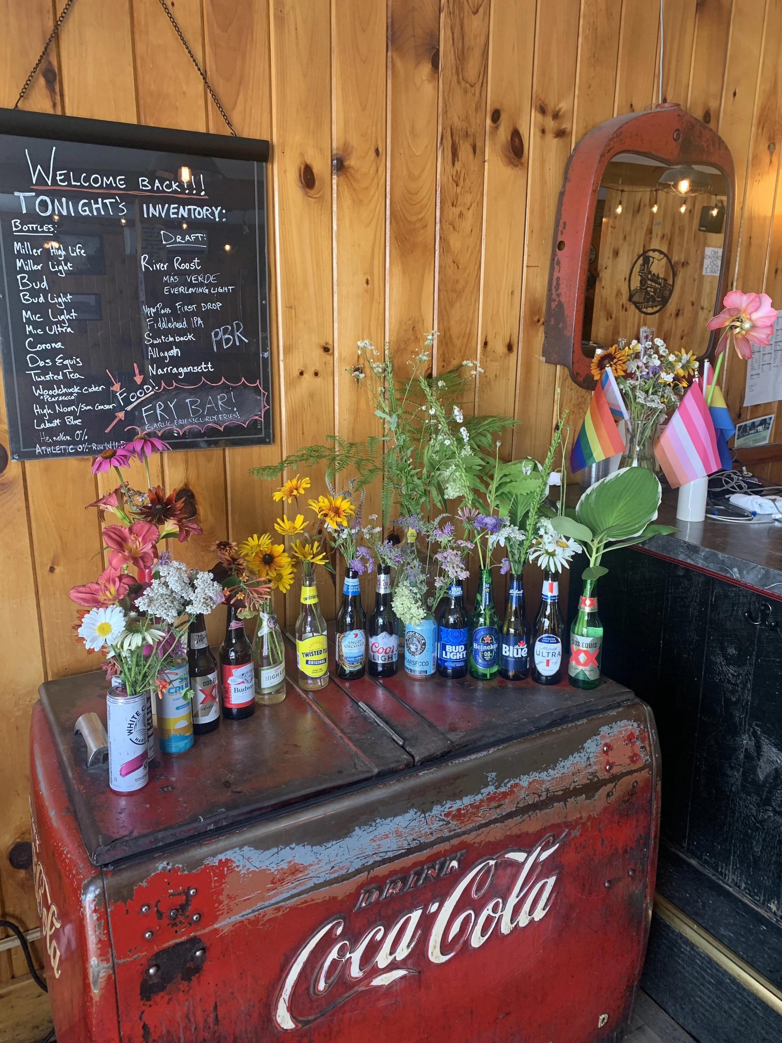 A vintage Coca-Cola cooler decorated with assorted colorful flowers in bottles, placed in a wooden-paneled interior with a chalkboard menu and small rainbow flags.