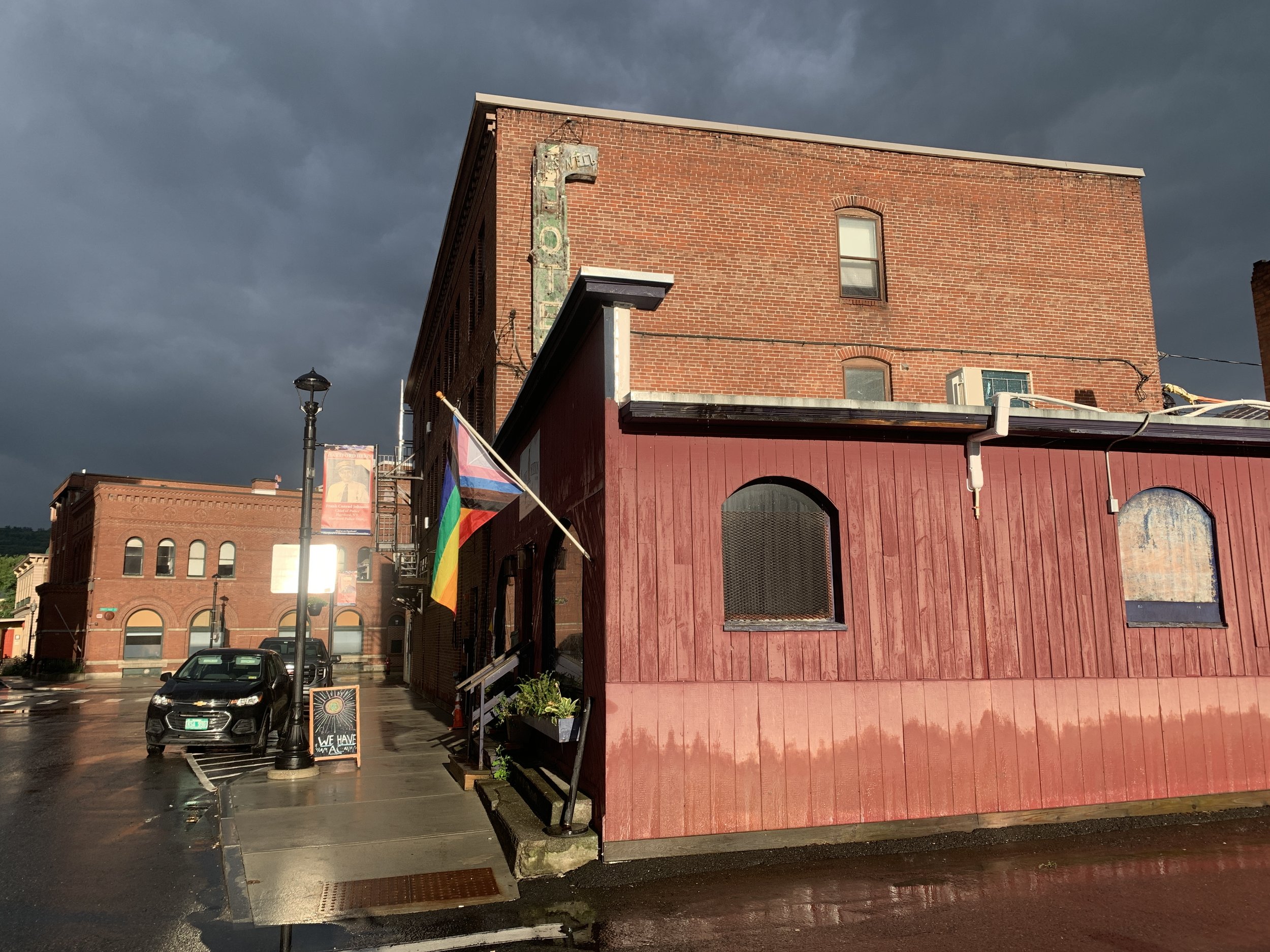 A street scene with a brick building, rainbow flag, and a parking lot on a rainy day with dark storm clouds overhead.