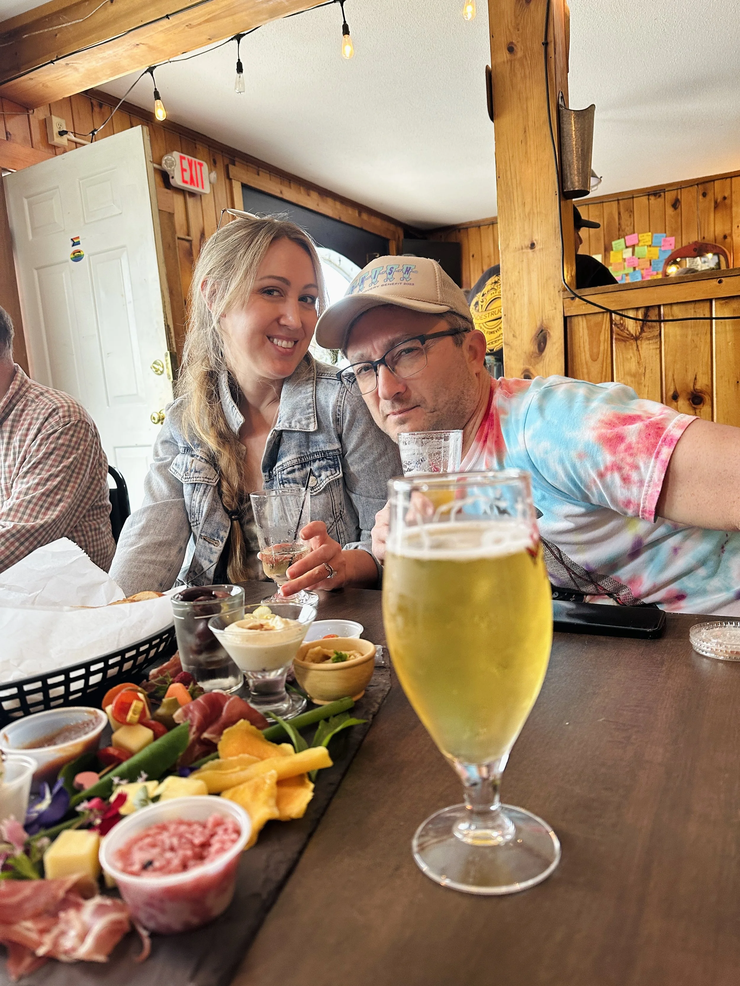 A woman and a man sitting at a table in a restaurant, with various food and drinks on the table, including a glass of beer, a glass of wine, and a cheese platter. The woman is smiling, and the man is holding the beer. The restaurant has wooden walls and ceiling, with string lights hanging above.