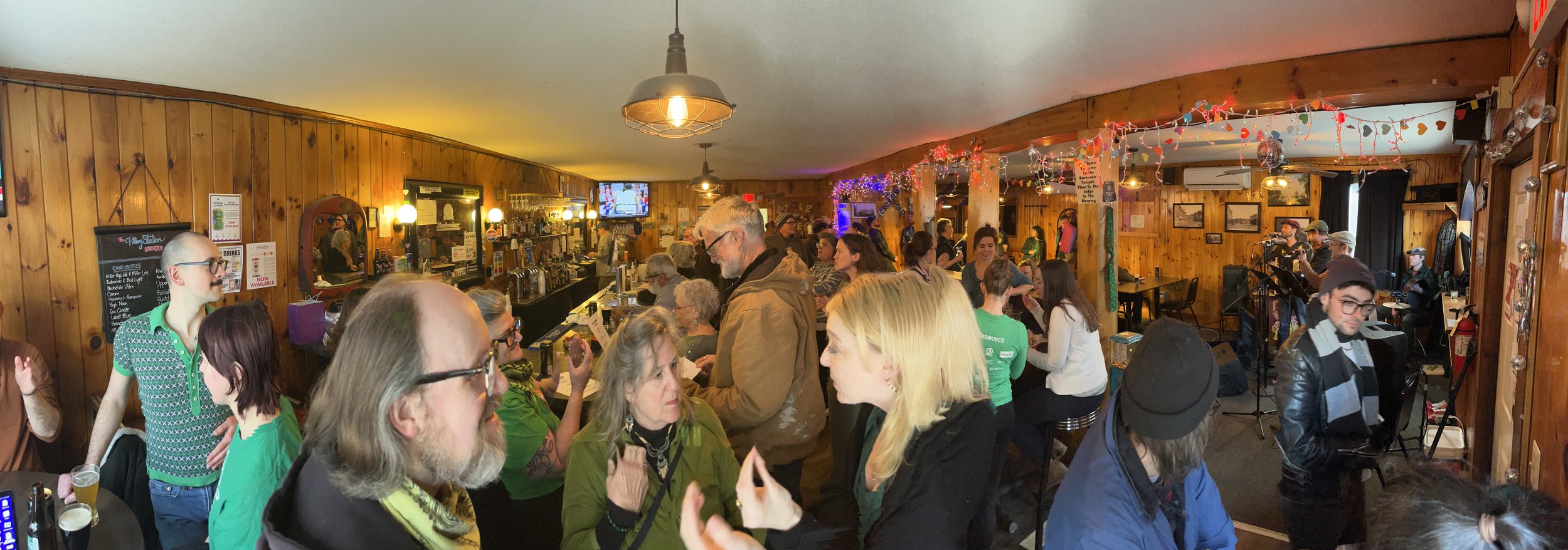 Crowded indoor bar or music venue with people socializing, a band performing on stage, wood-paneled walls, festive string lights, and a television screen above the bar.