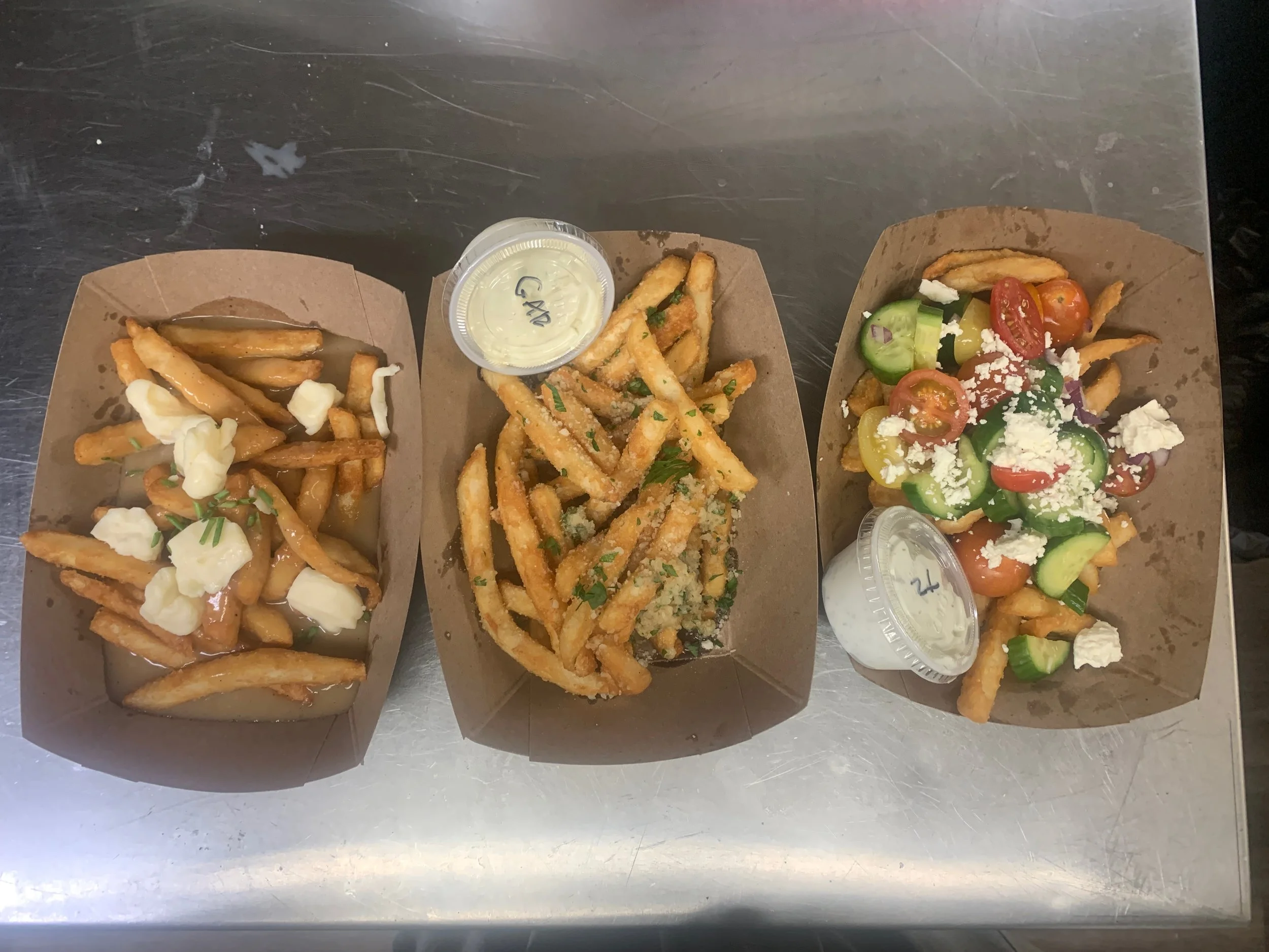 Three paper food trays with French fries topped with cheese, ranch dressing, and gravy, seasoned French fries with green herbs and cheese, and a loaded fry bowl with cherry and cucumber tomatoes, shredded cheese, and ranch dressing, on a metal surfac