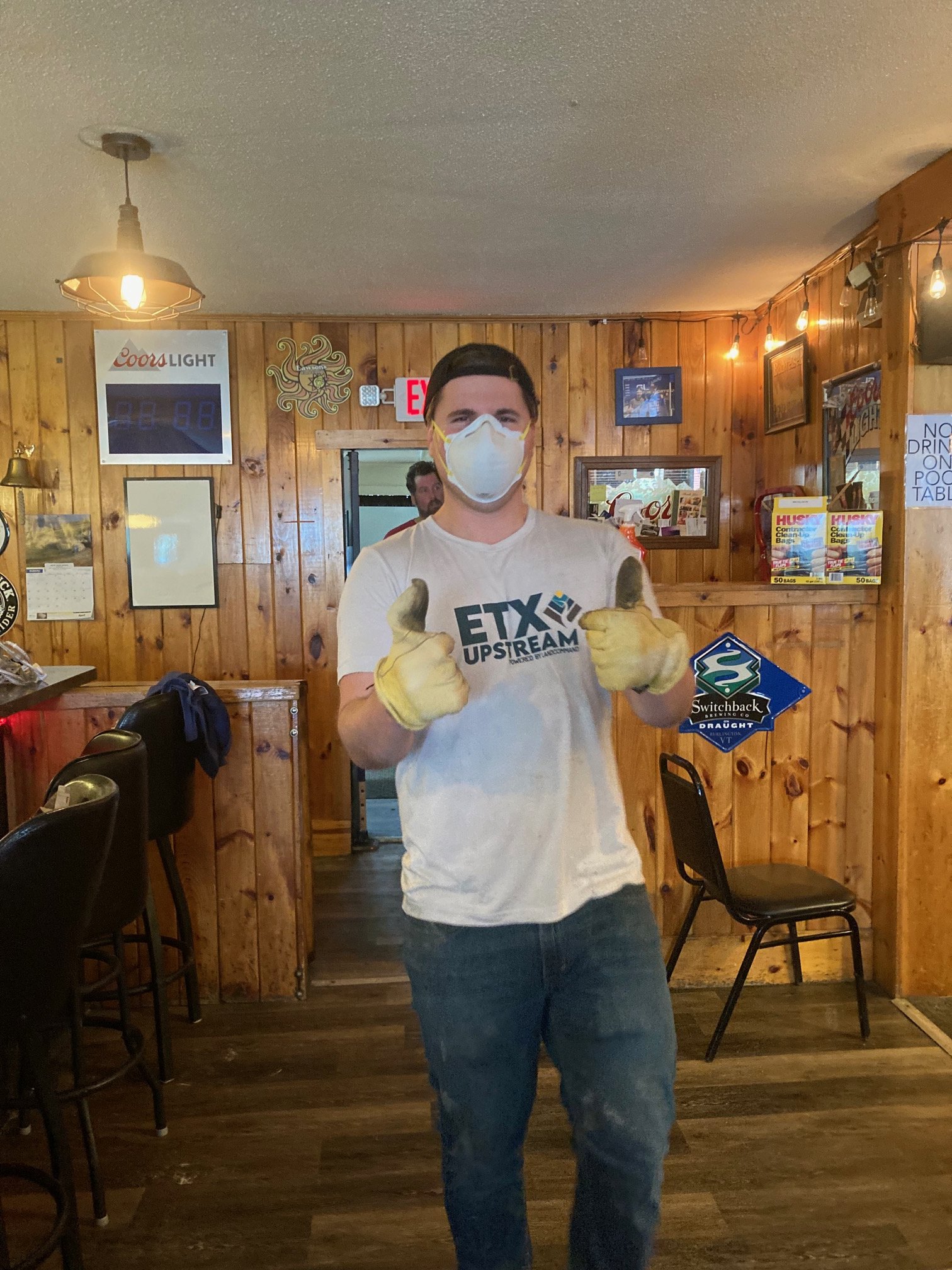 A young man wearing a face mask, yellow gloves, and a white T-shirt with the text 'ETX UPSTREAM' is standing in a wood-paneled room, giving two thumbs up.