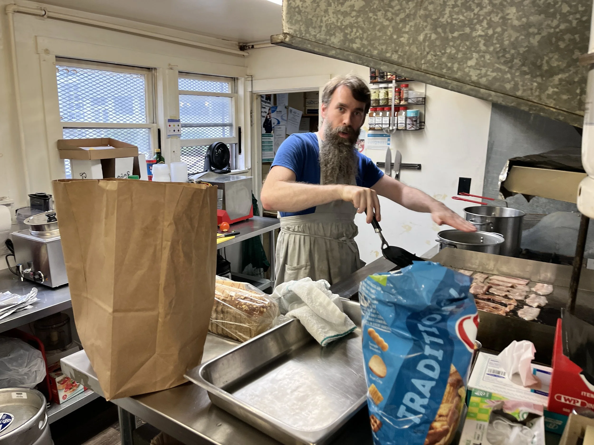 Man with a beard cooking bacon on a griddle in a commercial kitchen or food prep area.