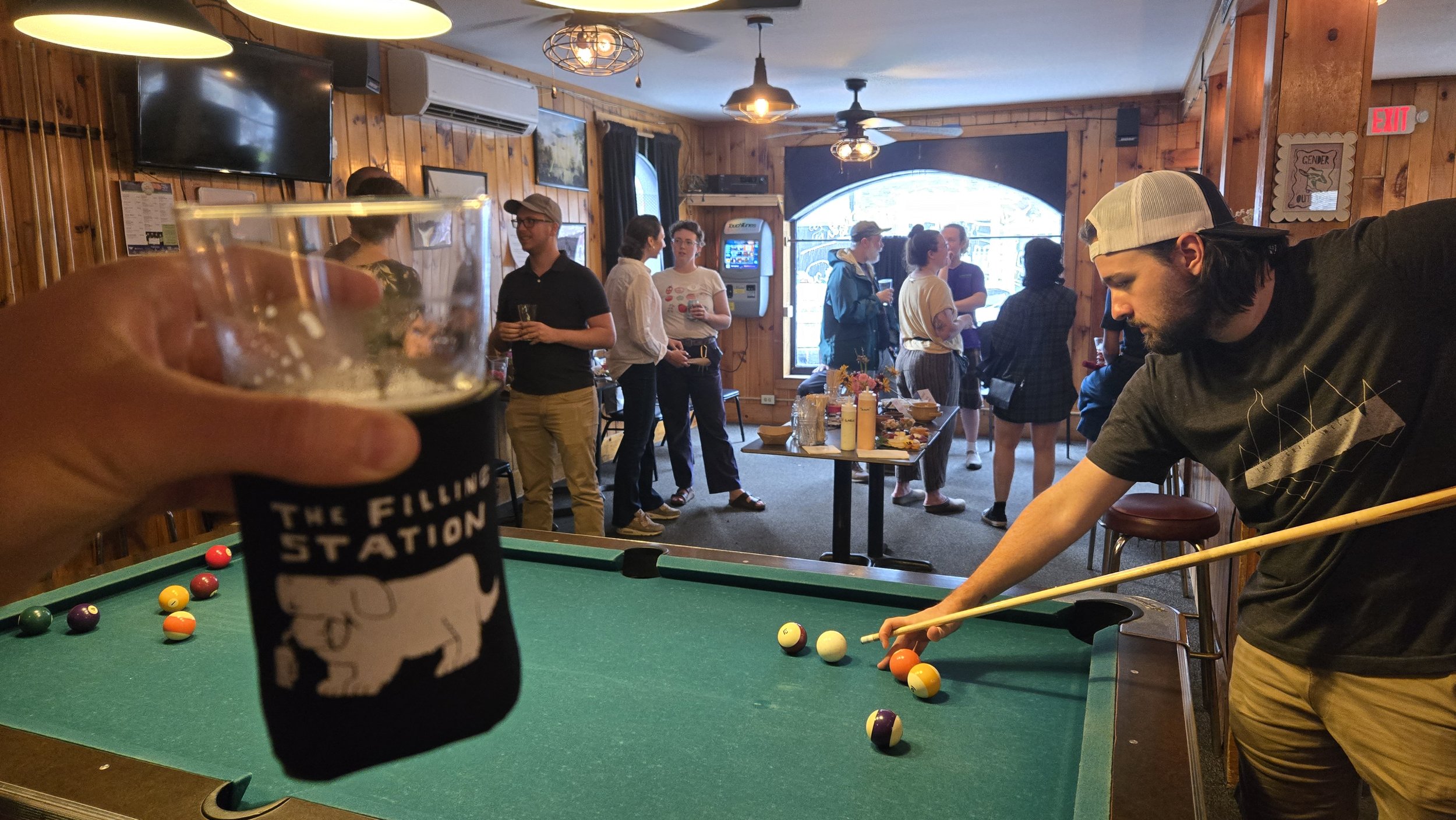 Person holding a cup in the foreground, with a group of people socializing in the background at a bar or social gathering, with a pool table in the scene.