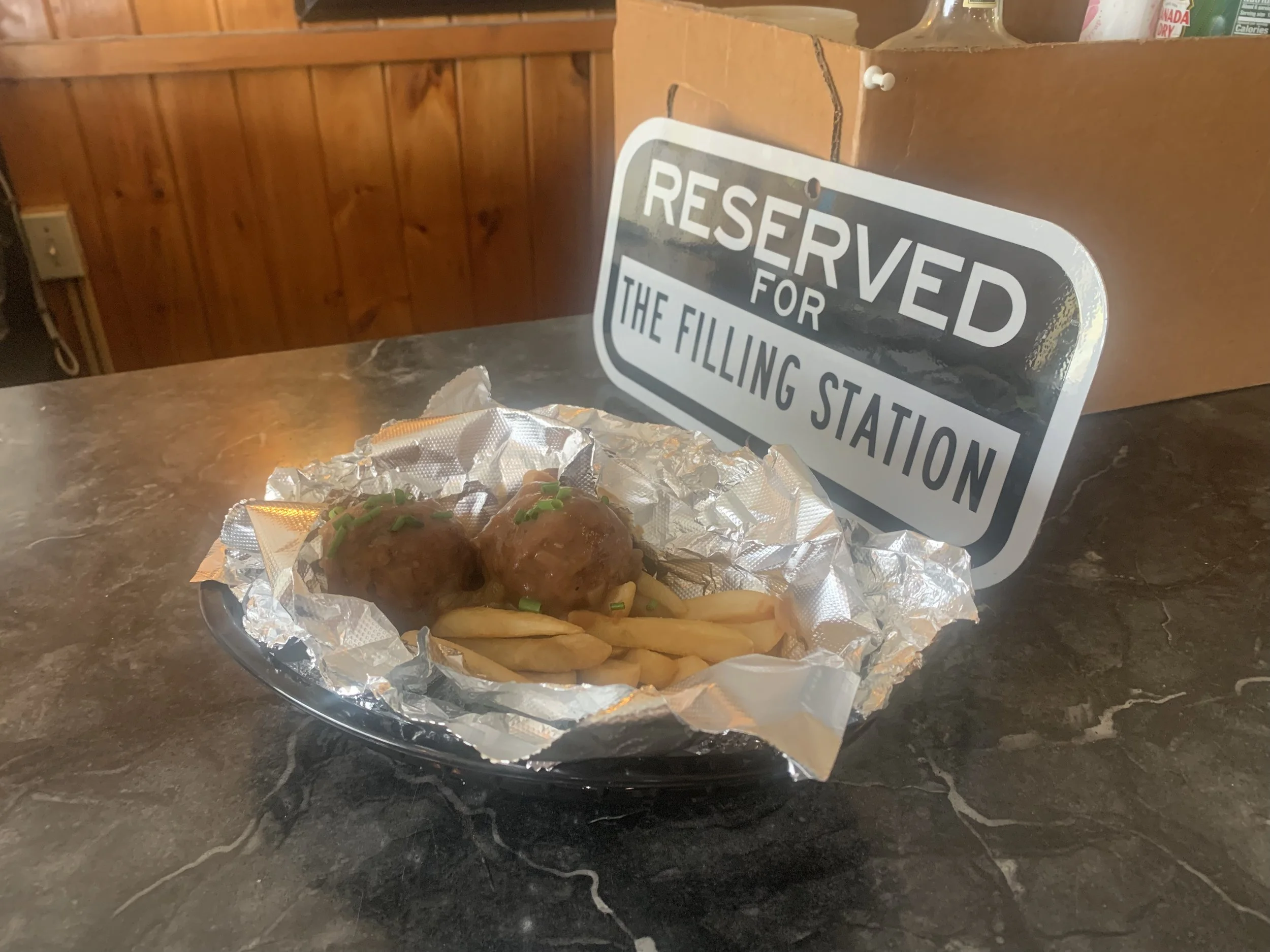 Plate with two meatballs, French fries, and chopped green onions, wrapped in aluminum foil, on a counter next to a reserve sign for the filling station.