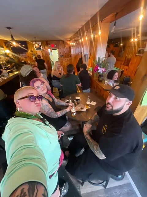 Selfie of four women and one man in a cozy restaurant or pub with wooden decor. The woman in the foreground is wearing glasses and a mint green hoodie, the others are seated at a table with drinks and a floral arrangement in the background.