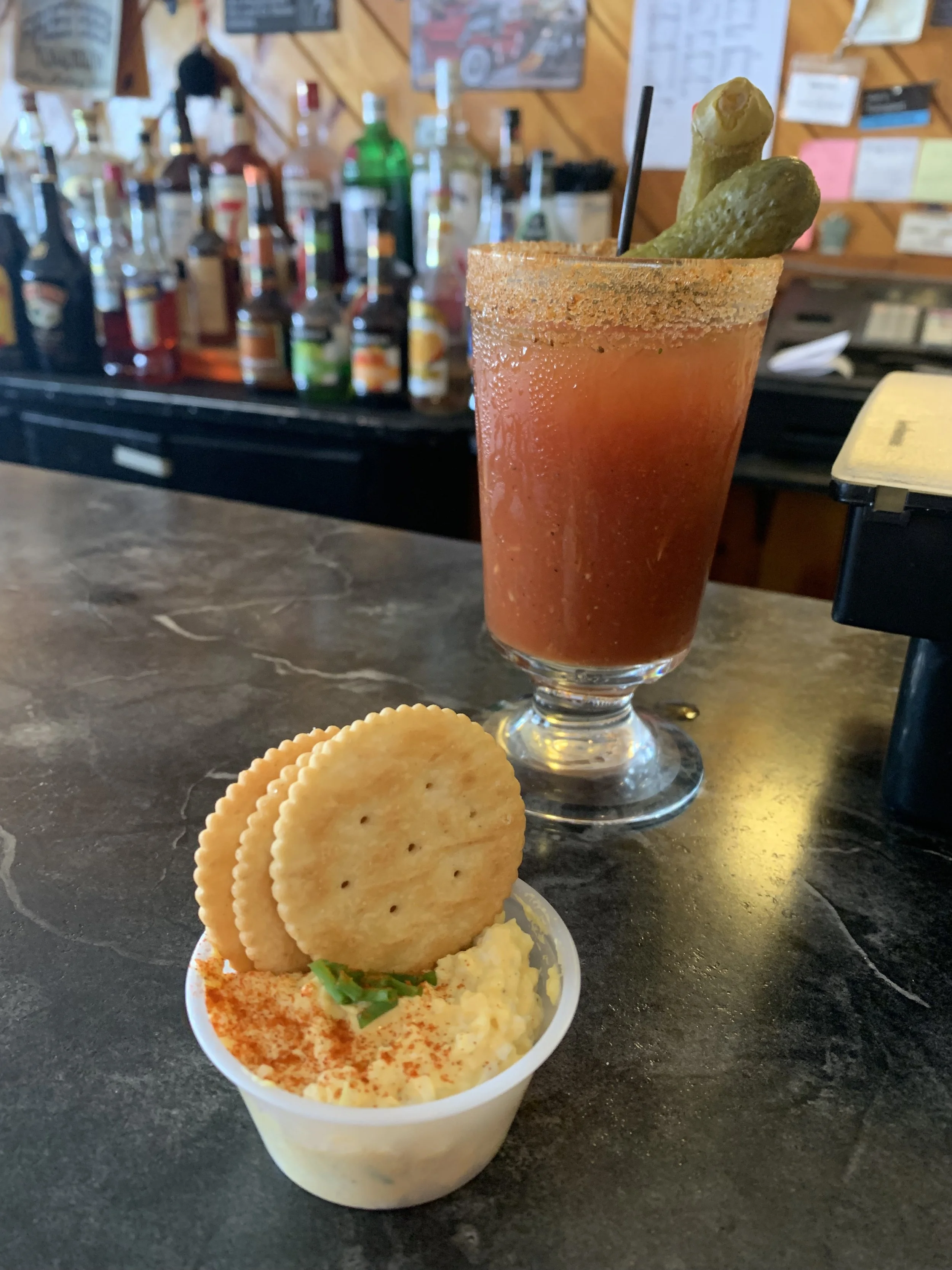 A Bloody Mary cocktail garnished with pickles on a straw, with salt or spice on the rim, sitting on a bar counter. Next to it, a small cup of potato chips and a scoop of potato salad garnished with paprika and a green onion.