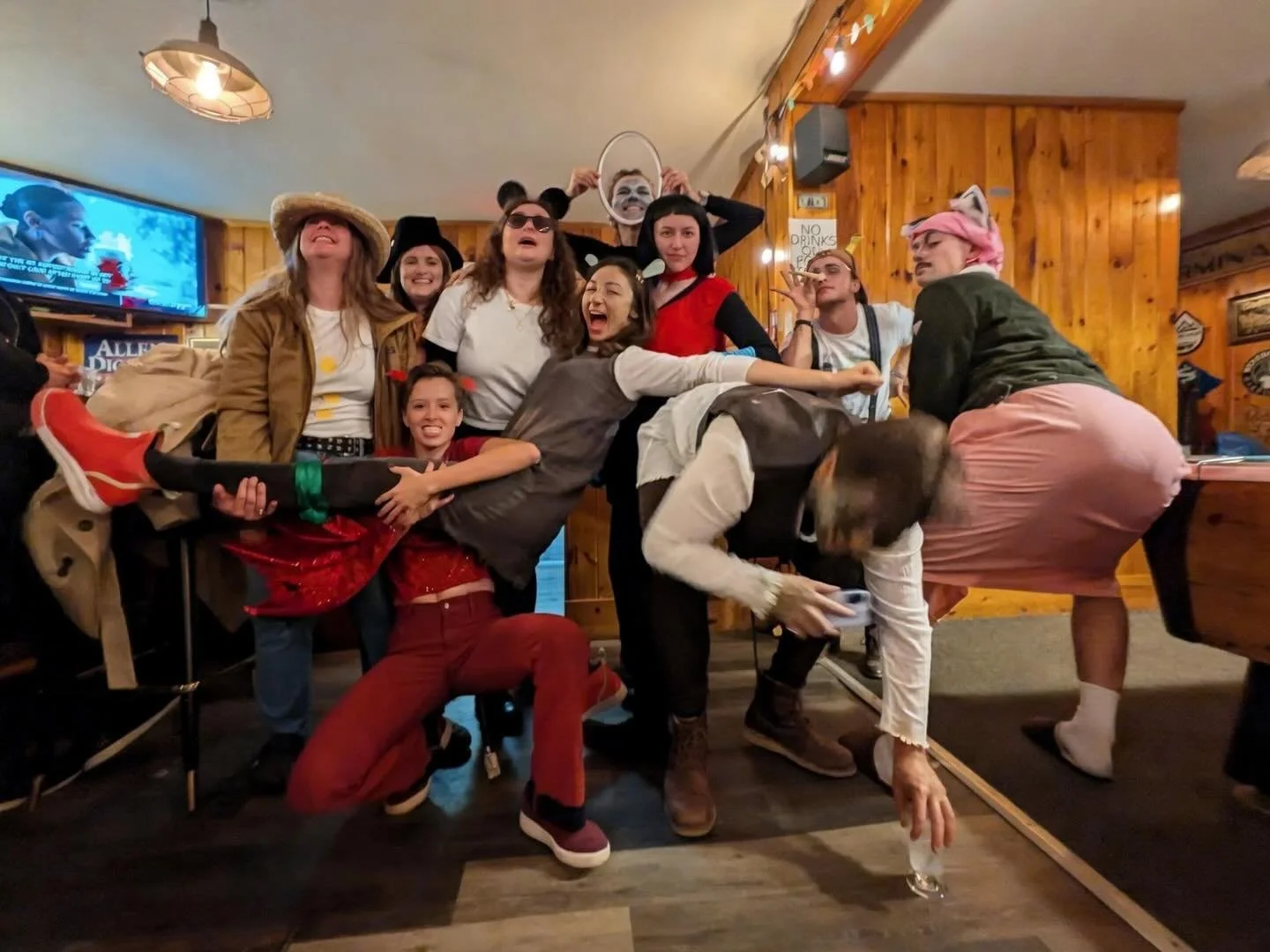 Group of people dressed in costumes at a party in a wooden-paneled room, some smiling and posing, others in playful, animal, and fantasy costumes.