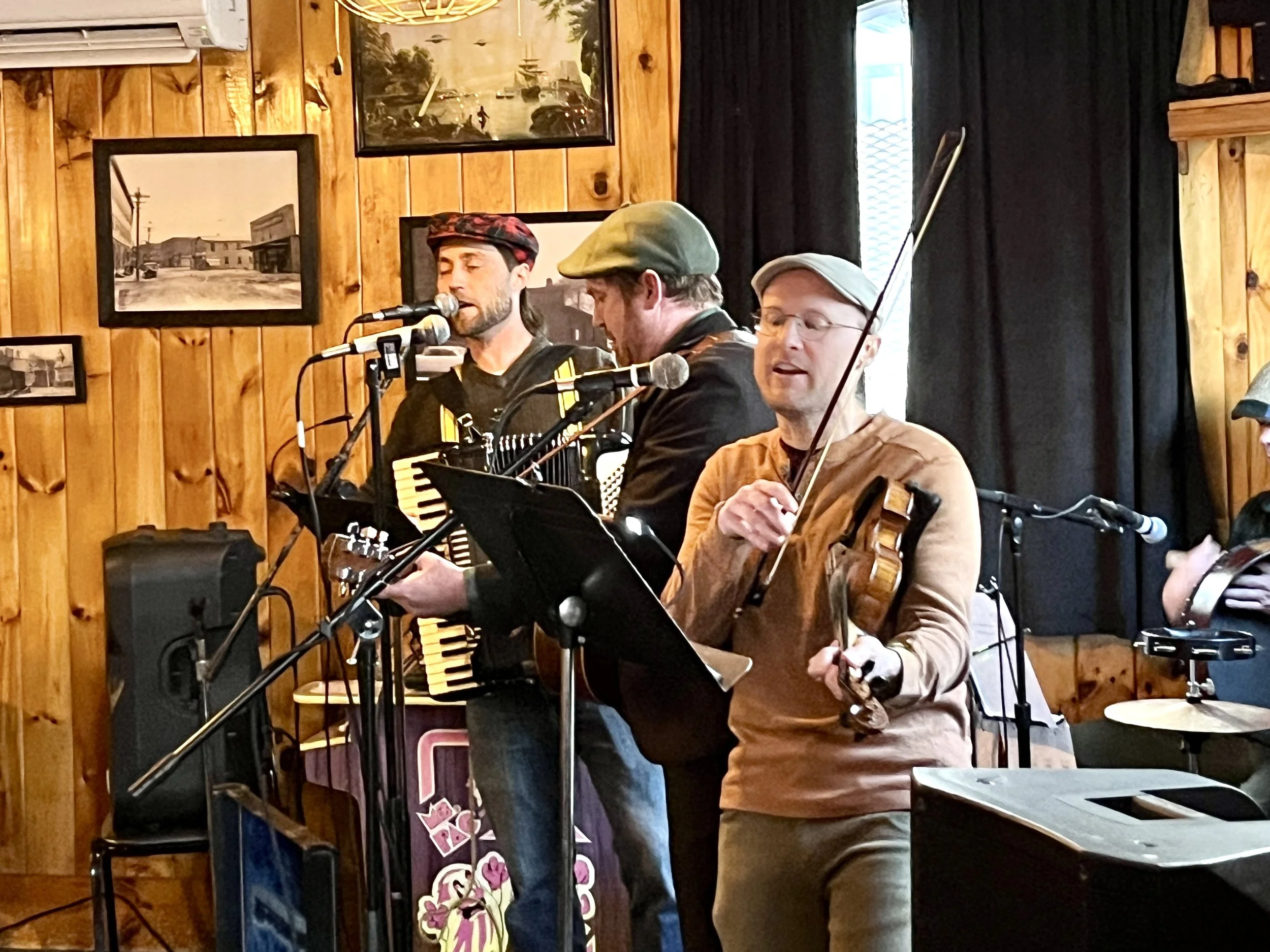 Three musicians performing indoors with wooden walls and framed pictures, one playing a violin, one with an accordion, and one singing into a microphone.