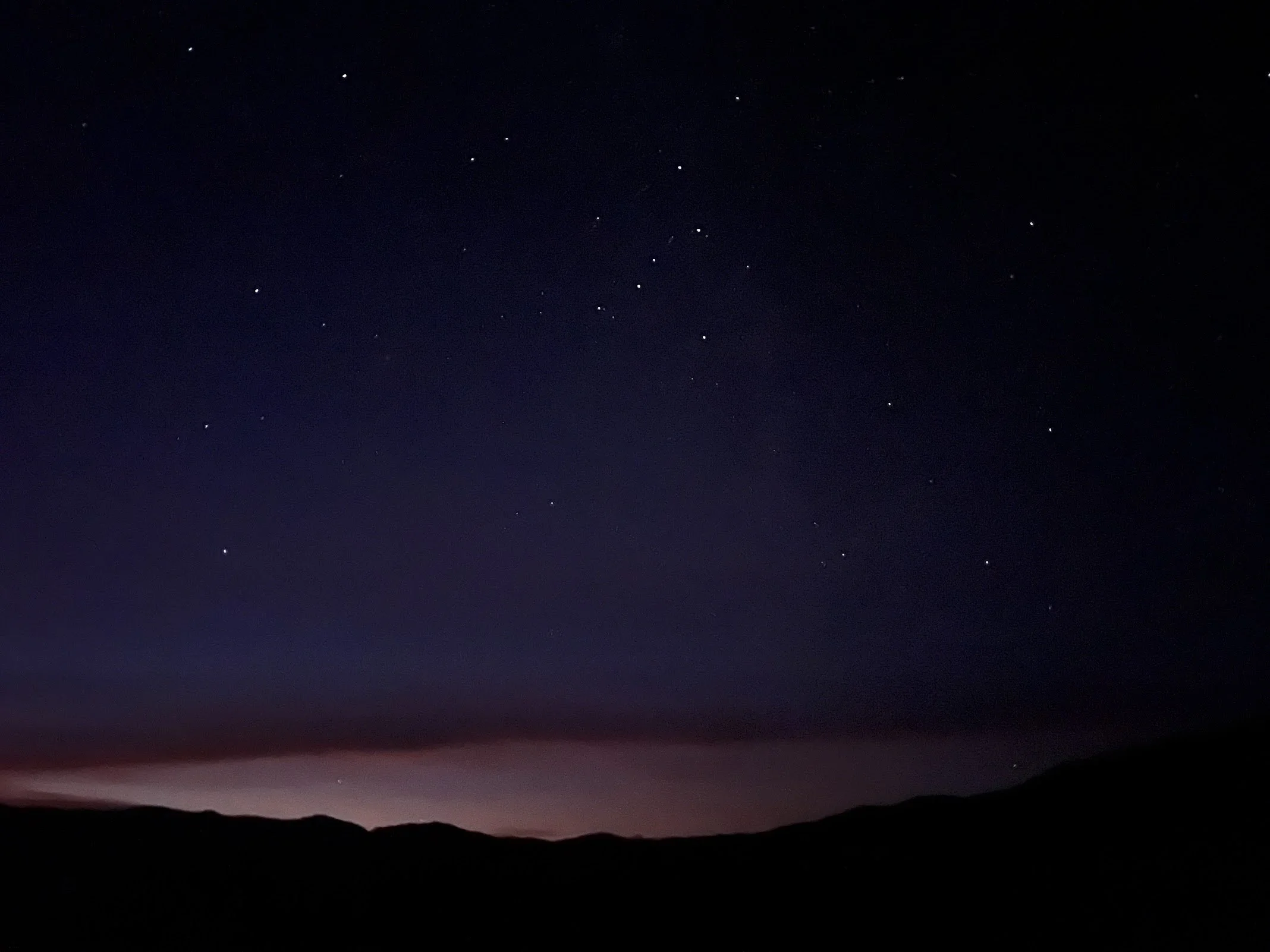 Night sky with numerous stars over dark mountains and a faint glow near the horizon.