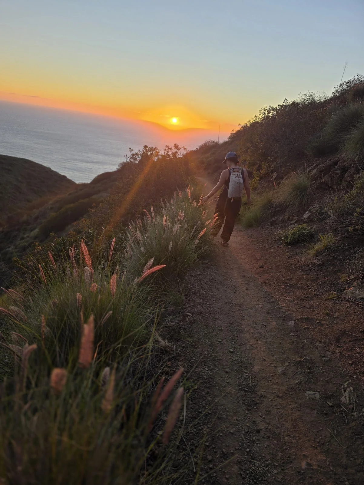 A person hiking on a trail along a hillside at sunset, with the ocean visible in the background and the sun near the horizon.