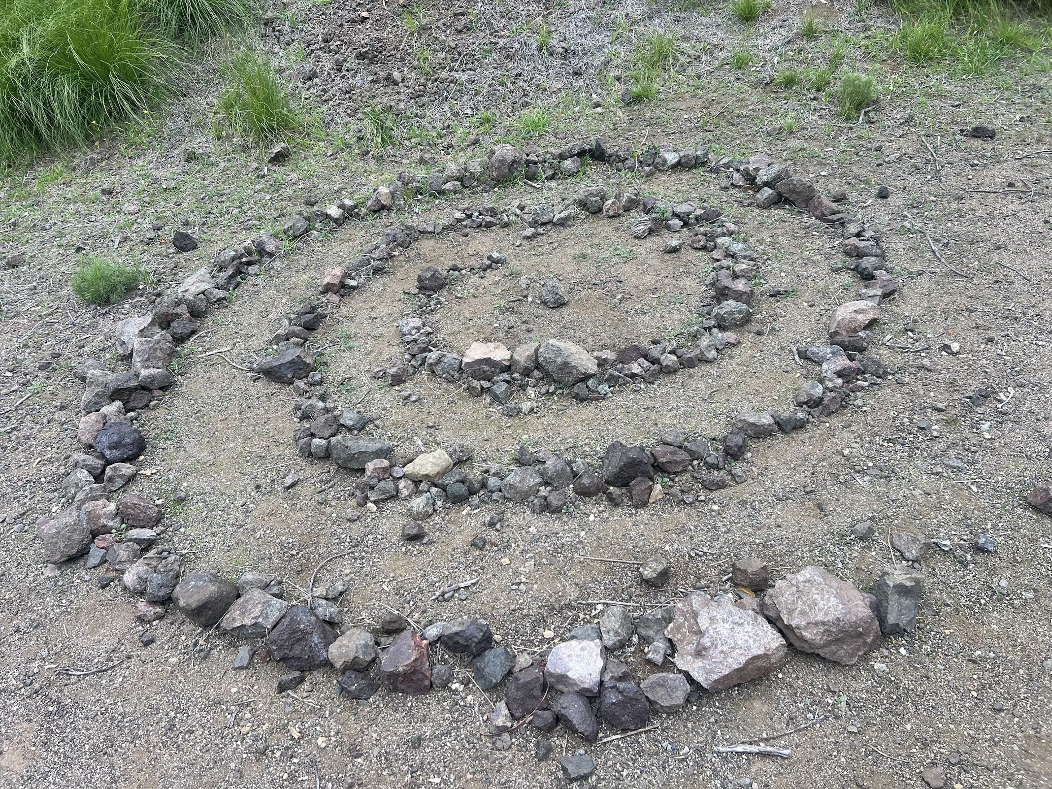 A spiral design made of small rocks and stones on the ground in an outdoor area with dirt and sparse grass.
