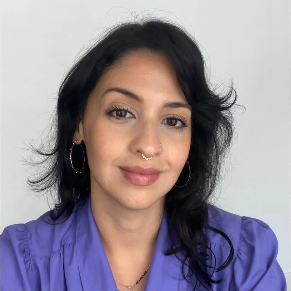 A woman with shoulder-length black hair, hoop earrings, a septum piercing, and a subtle smile, wearing a lavender blouse, standing against a plain white background.