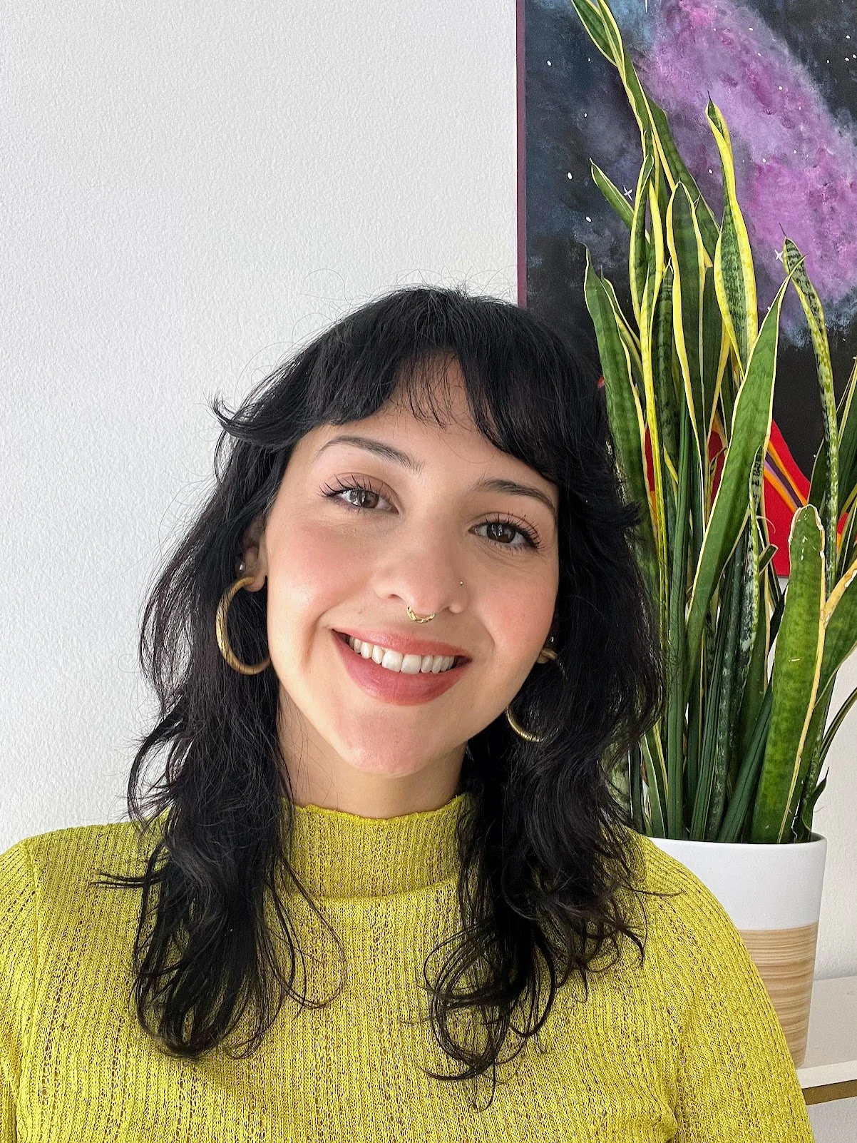 A woman with black wavy hair, wearing a yellow sweater, gold hoop earrings, a nose piercing, and a gold septum ring, smiling in front of a large potted plant with long green leaves and a colorful space-themed poster on the wall.